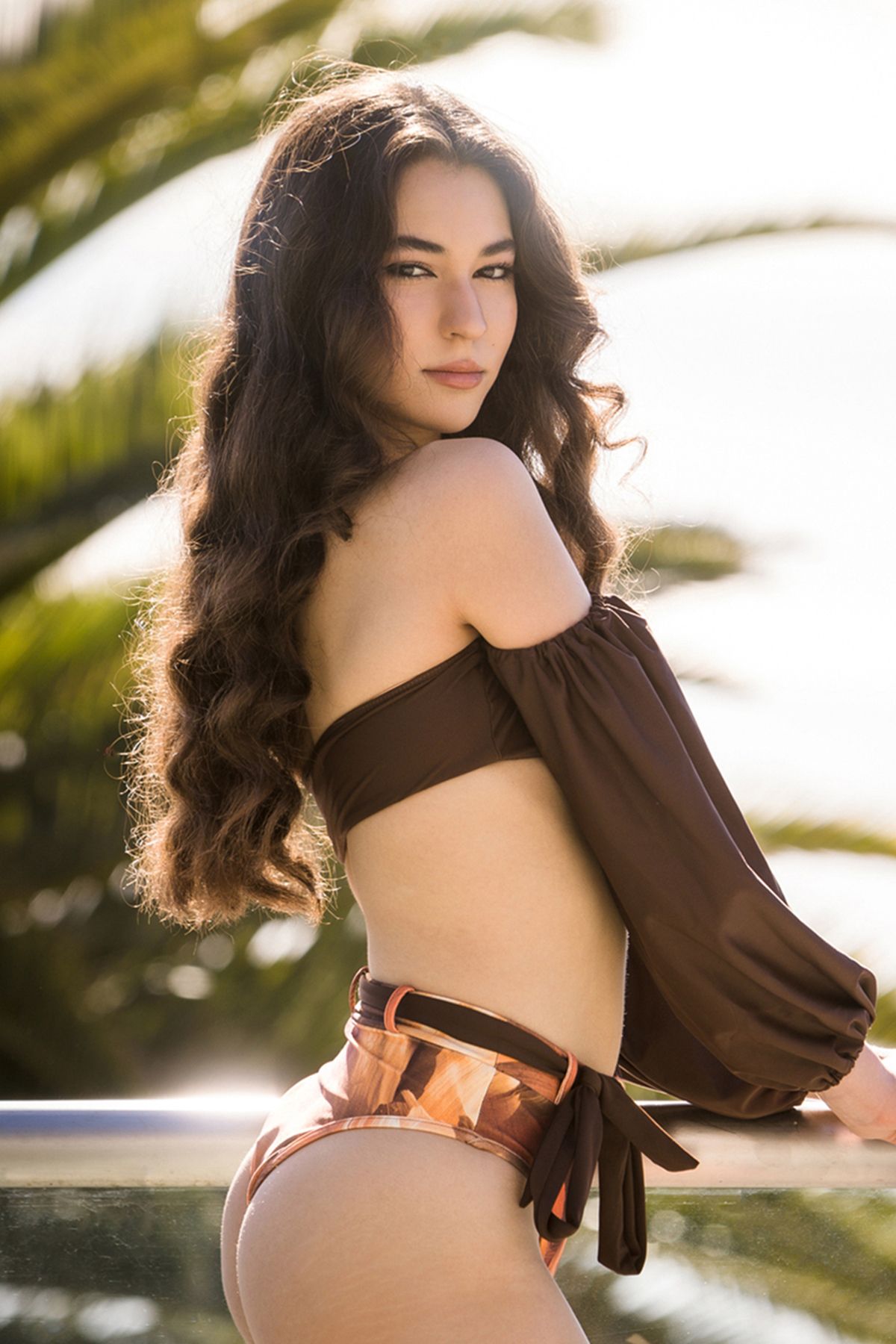 Young woman in brown off-shoulder crop top and metallic bikini bottoms posing outdoors near pool with palm trees.