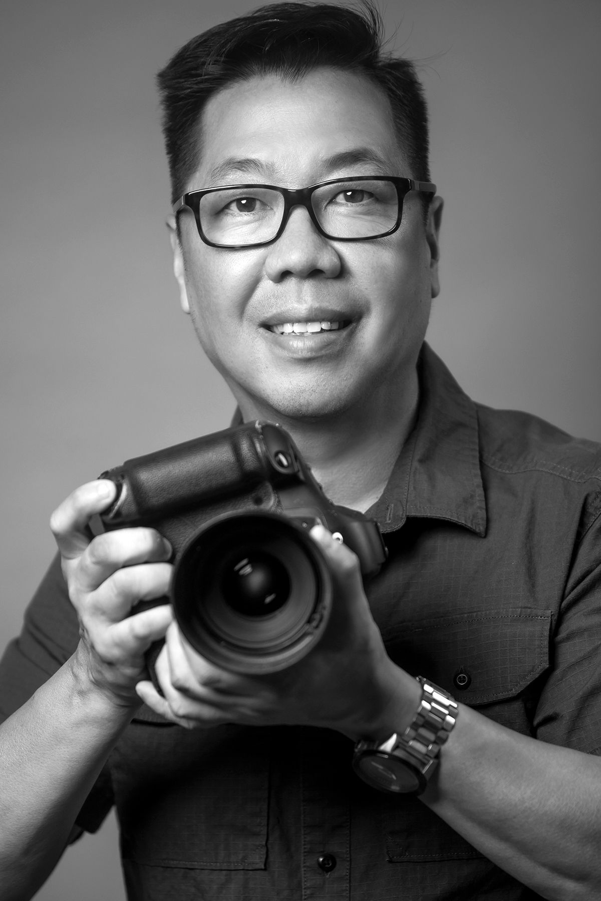 Professional photographer smiling while holding a DSLR camera in a black and white studio portrait.