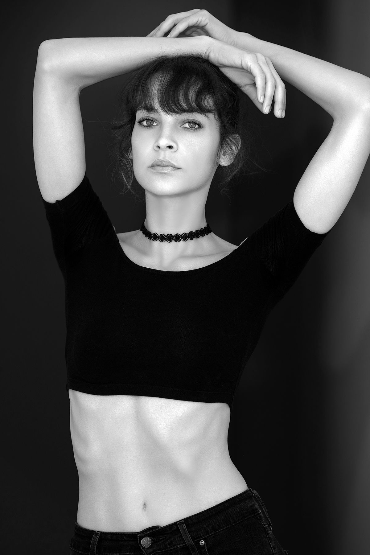 Black and white portrait of a woman in a crop top with bangs, arms raised above her head.