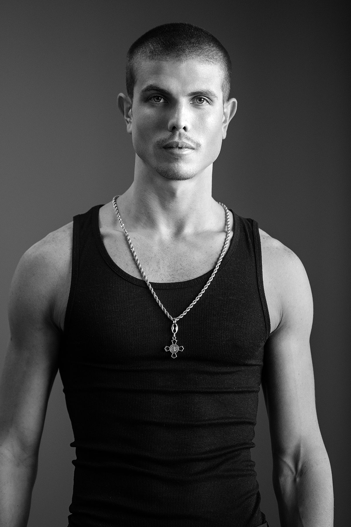 Black and white portrait of a muscular young man in a tank top wearing a cross chain necklace.