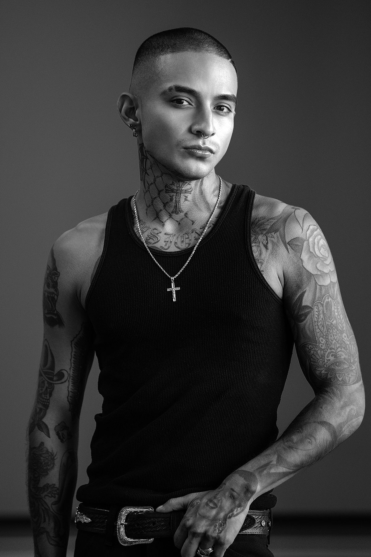 Heavily tattooed man with shaved head in black tank top and cross necklace in studio portrait.