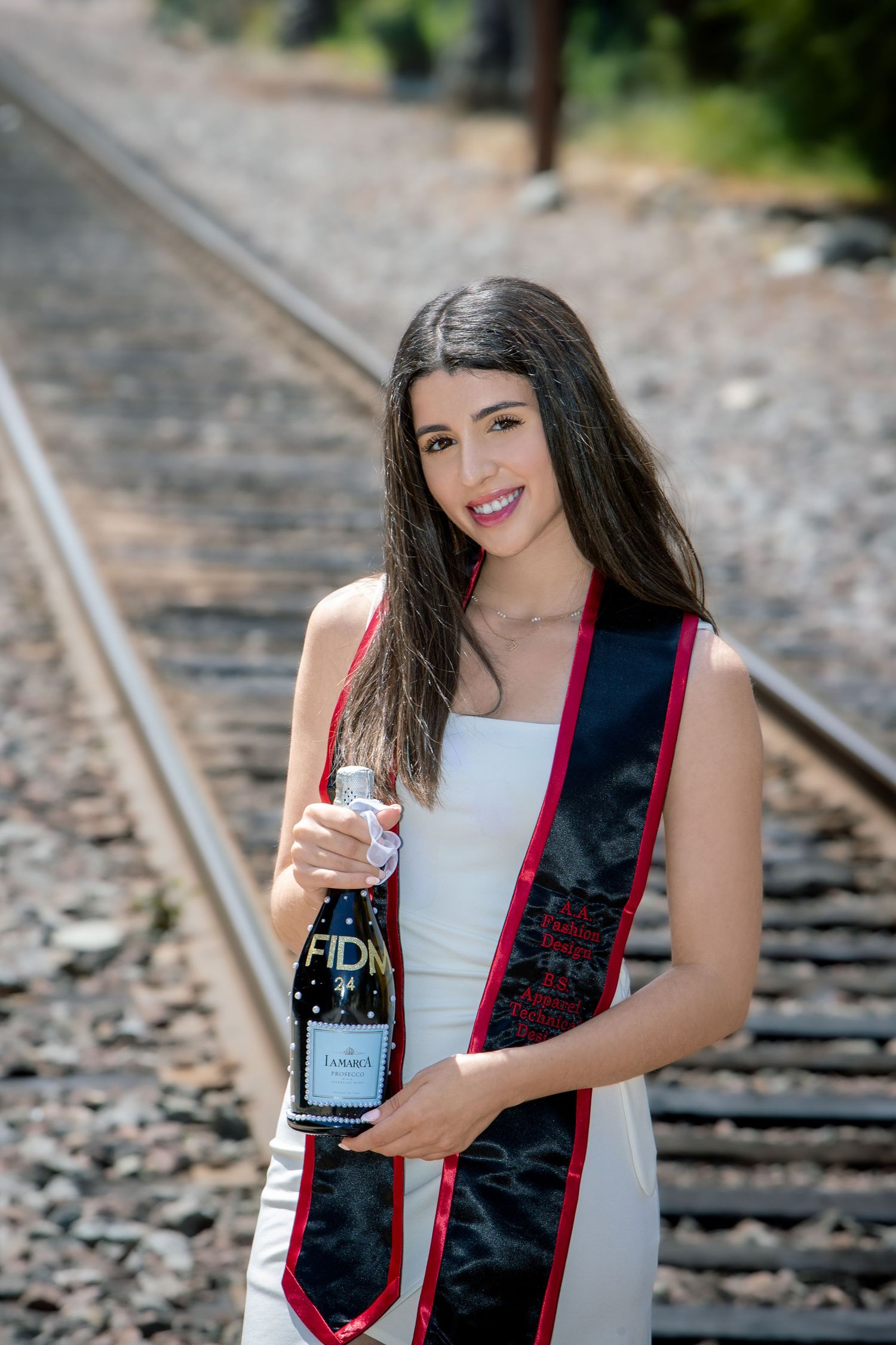 Female FIDM graduate in white dress holds champagne bottle on railroad tracks during outdoor graduation photo session.