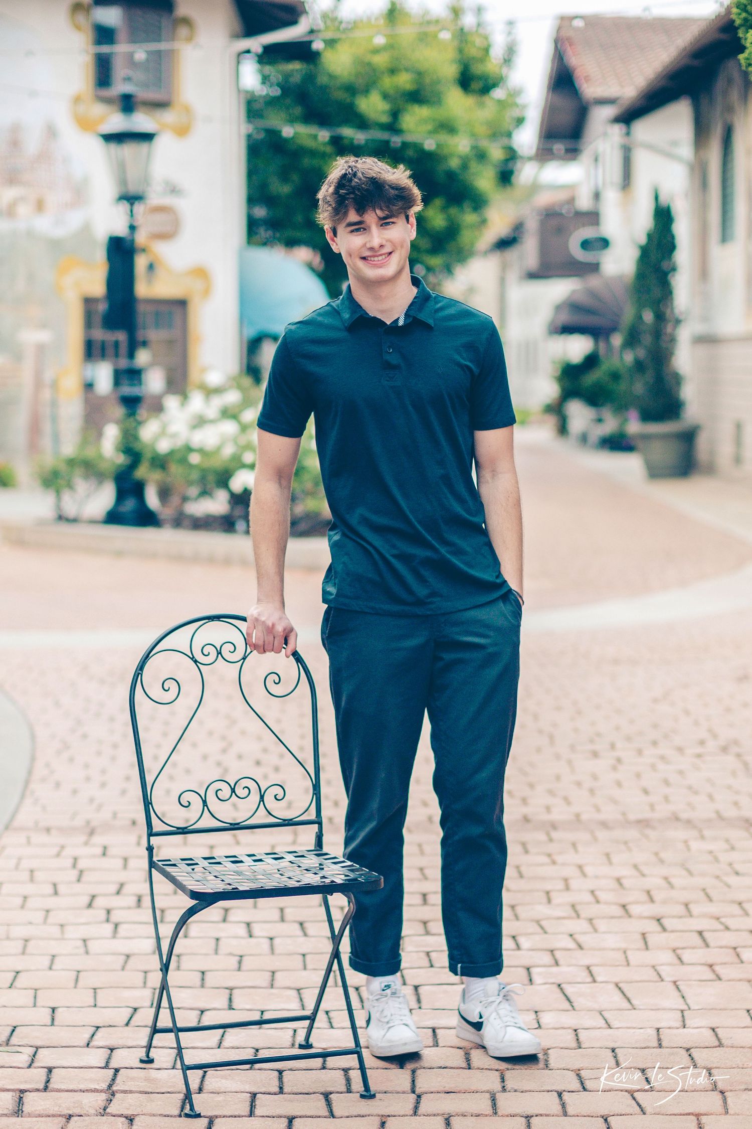 Tall young man in dark teal polo standing beside an ornate iron chair on a brick plaza.