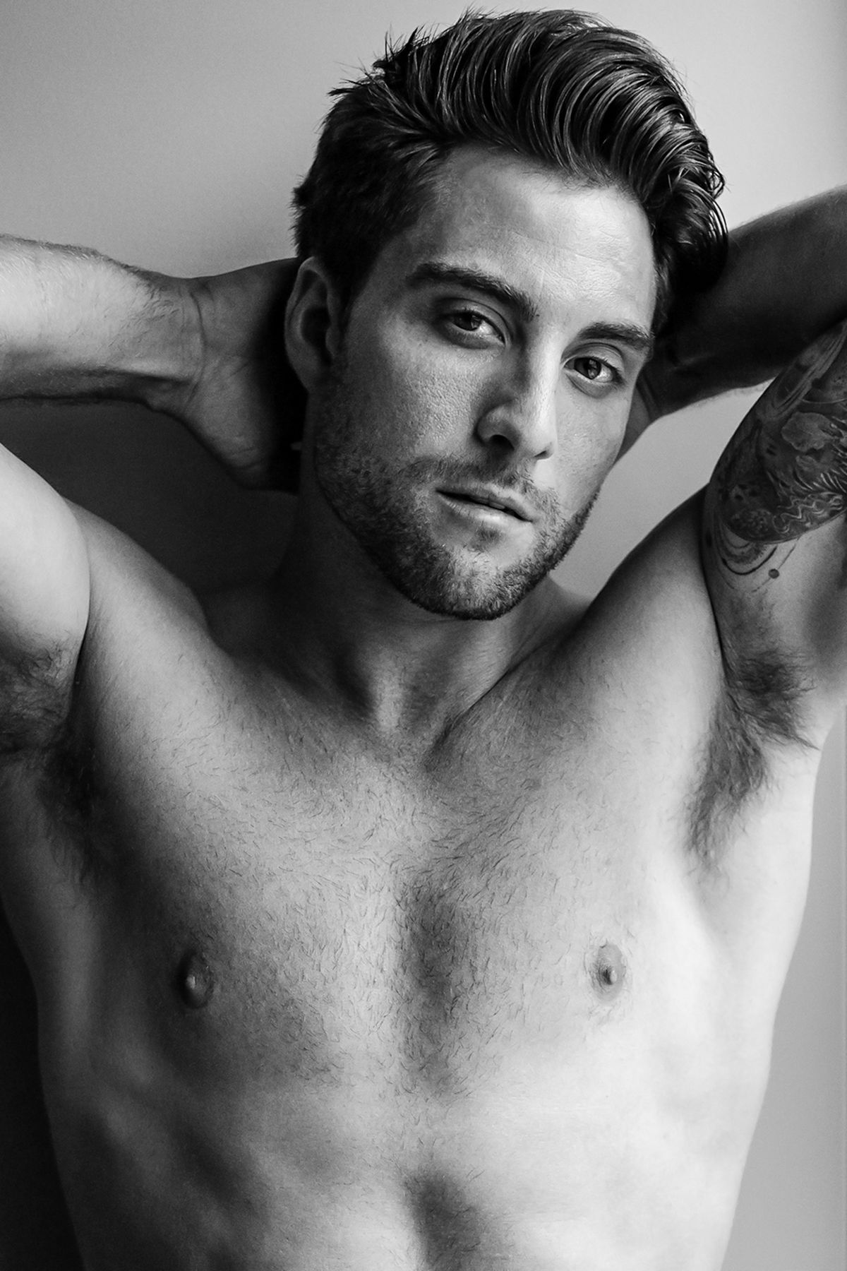 Shirtless muscular man with tattoo poses with hands behind head in black and white portrait photo.