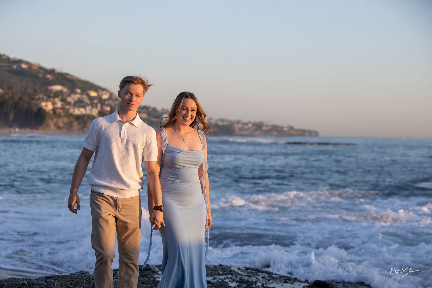 Couple walking hand in hand on rocky beach at sunset with ocean waves behind them.