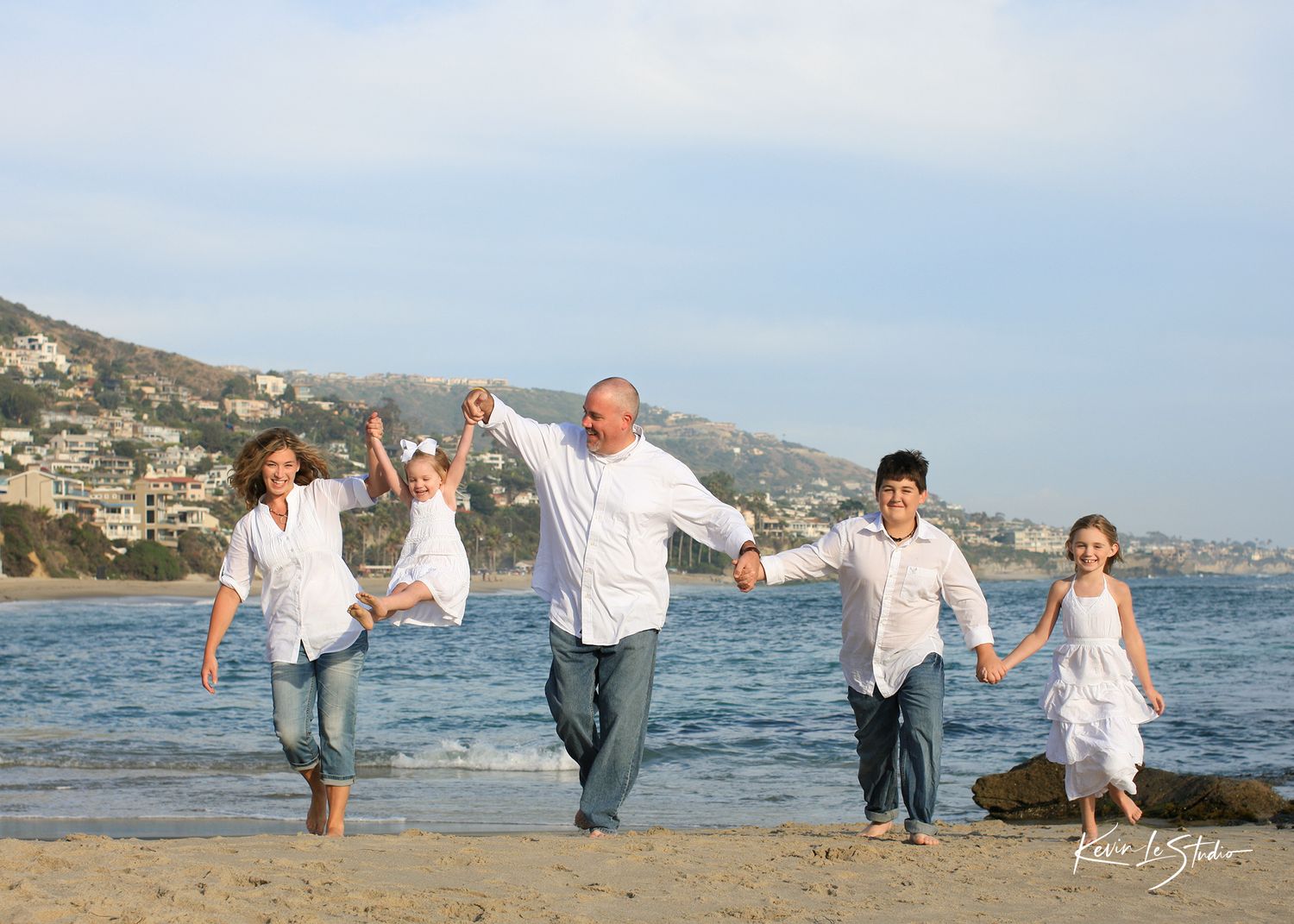 Happy family of five running and holding hands on a sunny beach with coastal hills in the background.