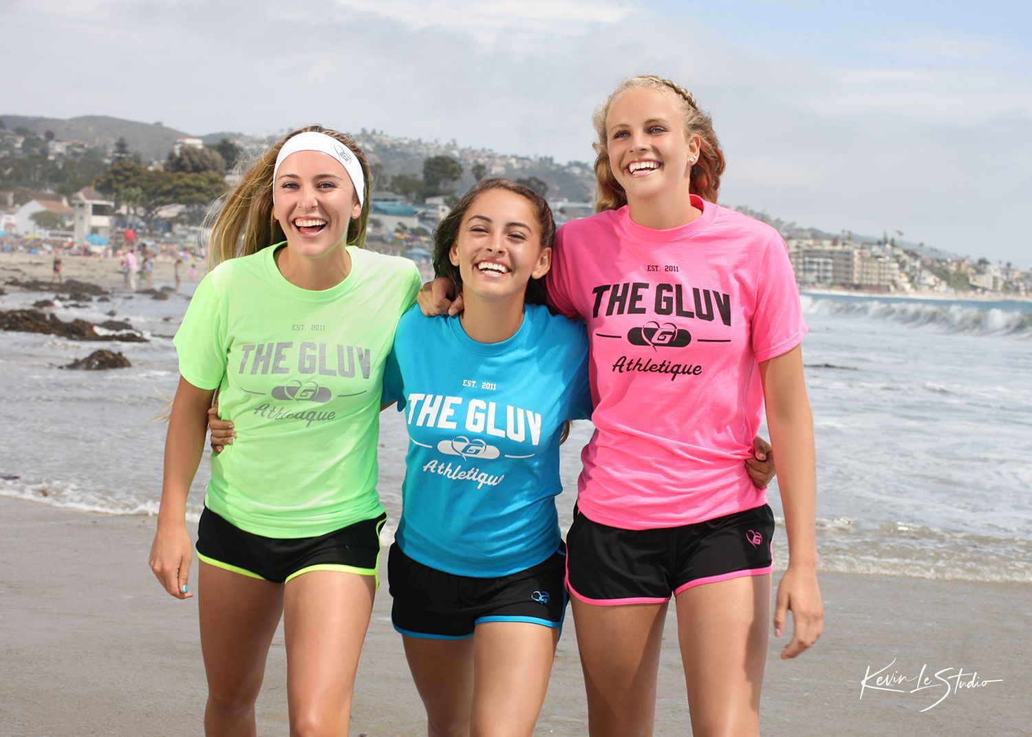 Three smiling girls wearing The Gluv Athletics t-shirts pose together on a sunny beach.