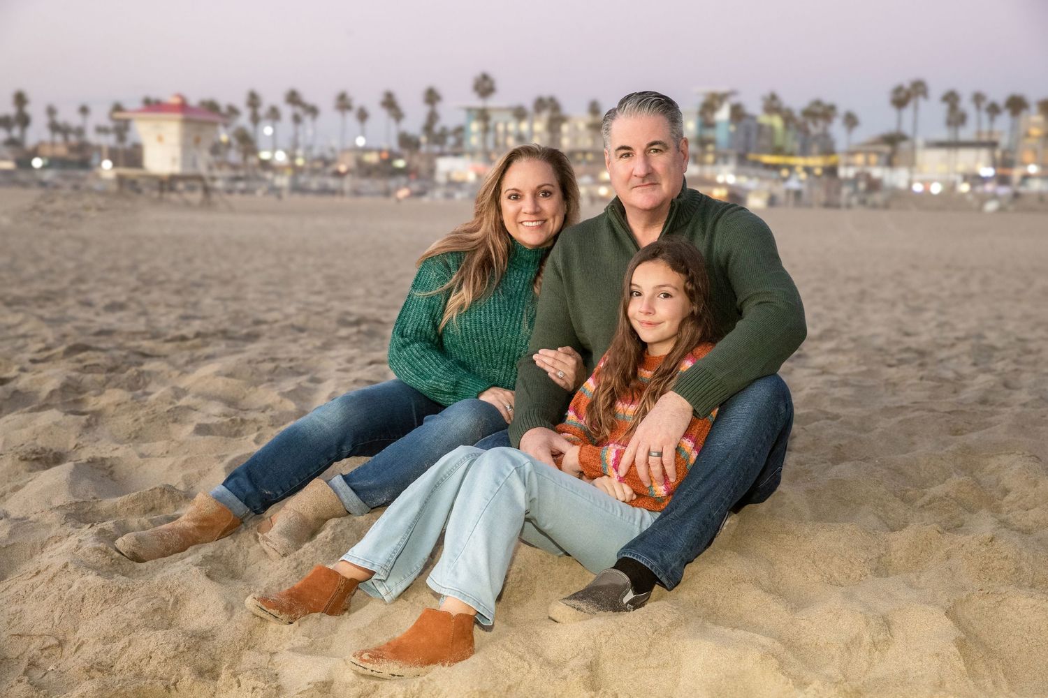 Family of three smiling together on a sandy beach at sunset, wearing green and casual outfits with palm trees in background.