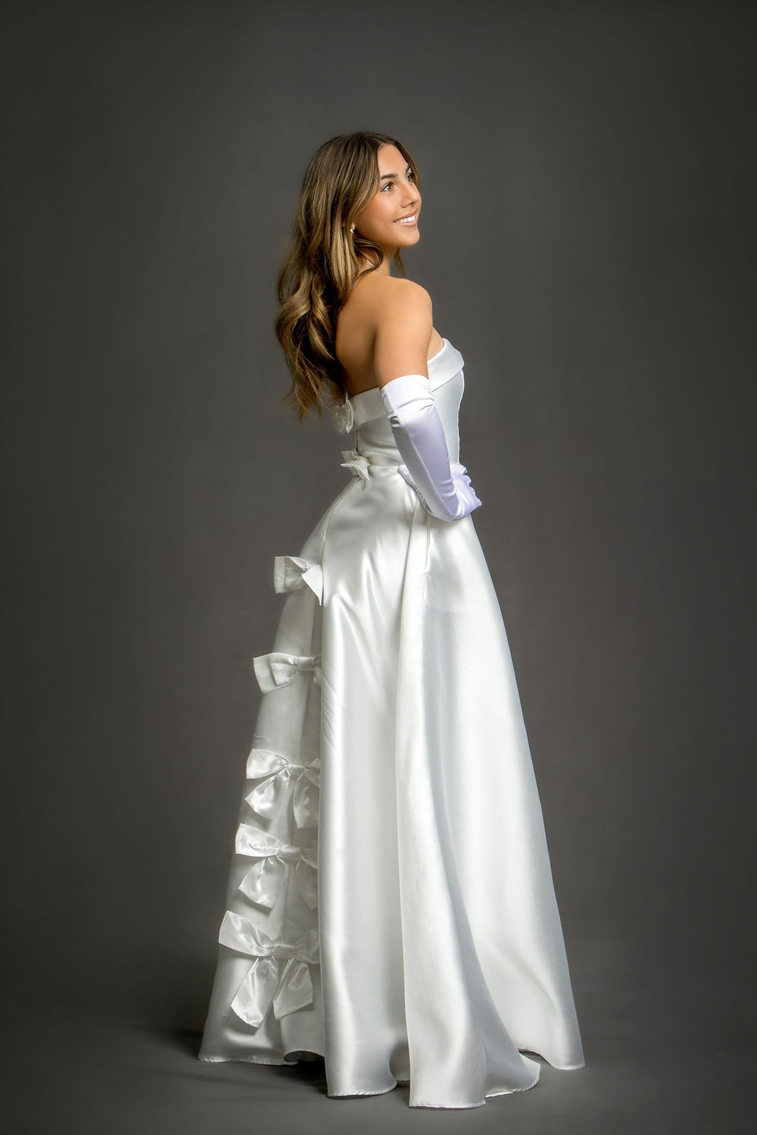 Smiling bride in elegant white satin strapless gown with ruffled skirt and long gloves, posed against gray background.