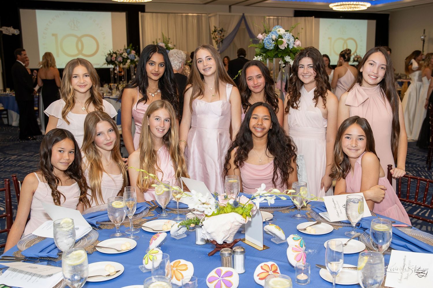 Group of young girls in elegant pink and white dresses posing together at a formal banquet table.