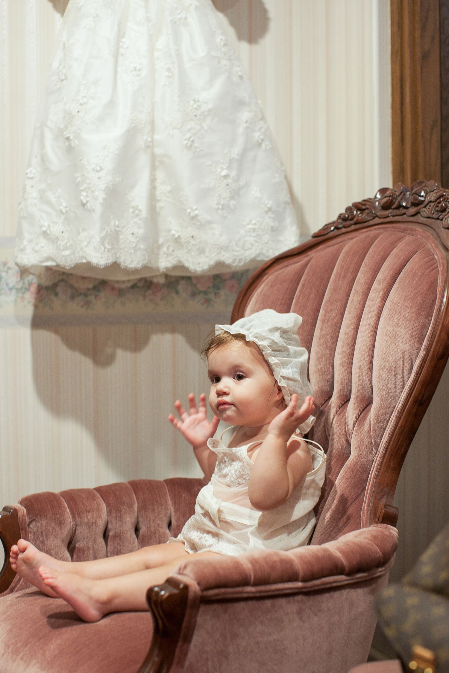 Adorable baby girl in white dress and bonnet sitting in a vintage pink velvet armchair, clapping hands.