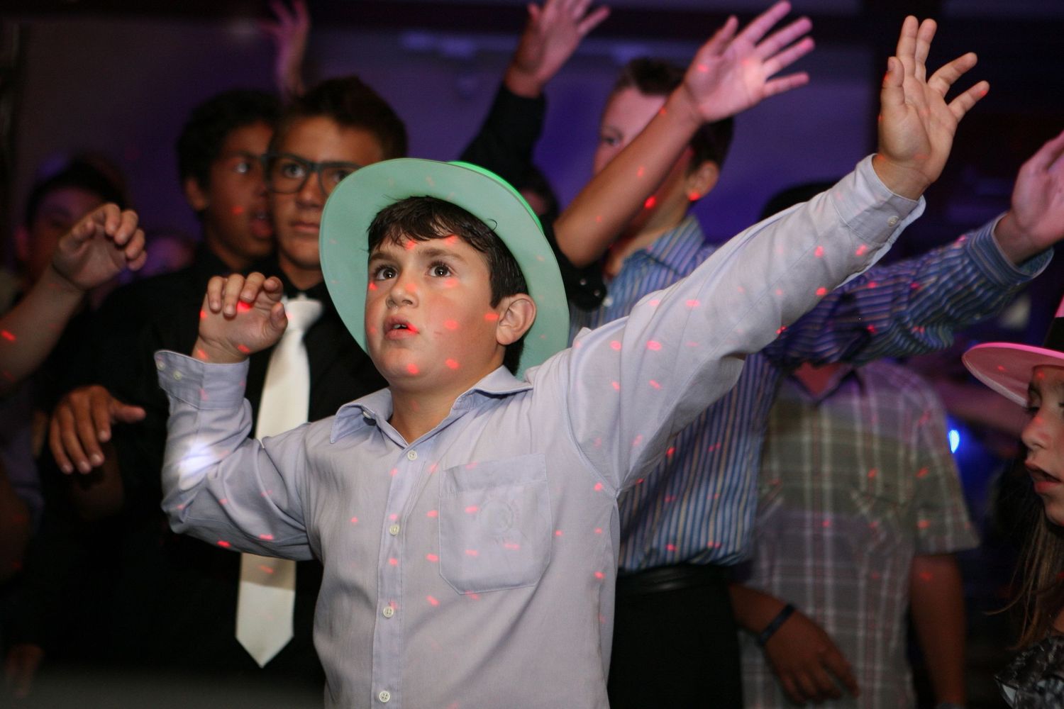 Young boy in light blue shirt and mint hat dancing with arms outstretched at a lively party celebration.