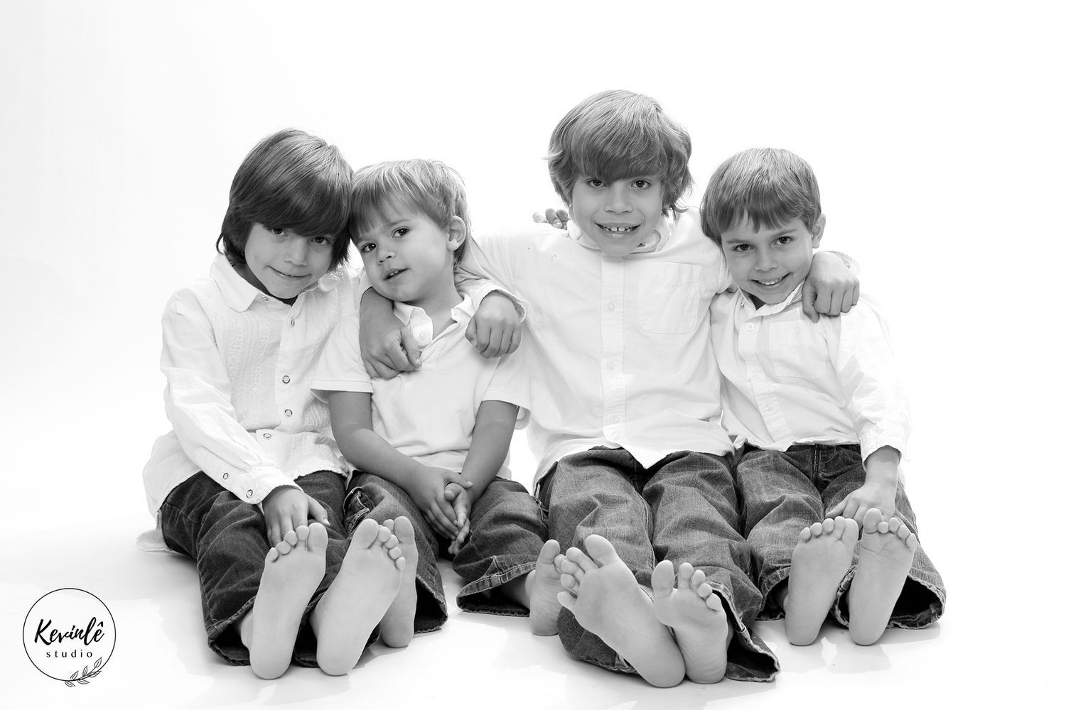 Black and white studio portrait of four young boys sitting together barefoot, smiling and hugging each other.