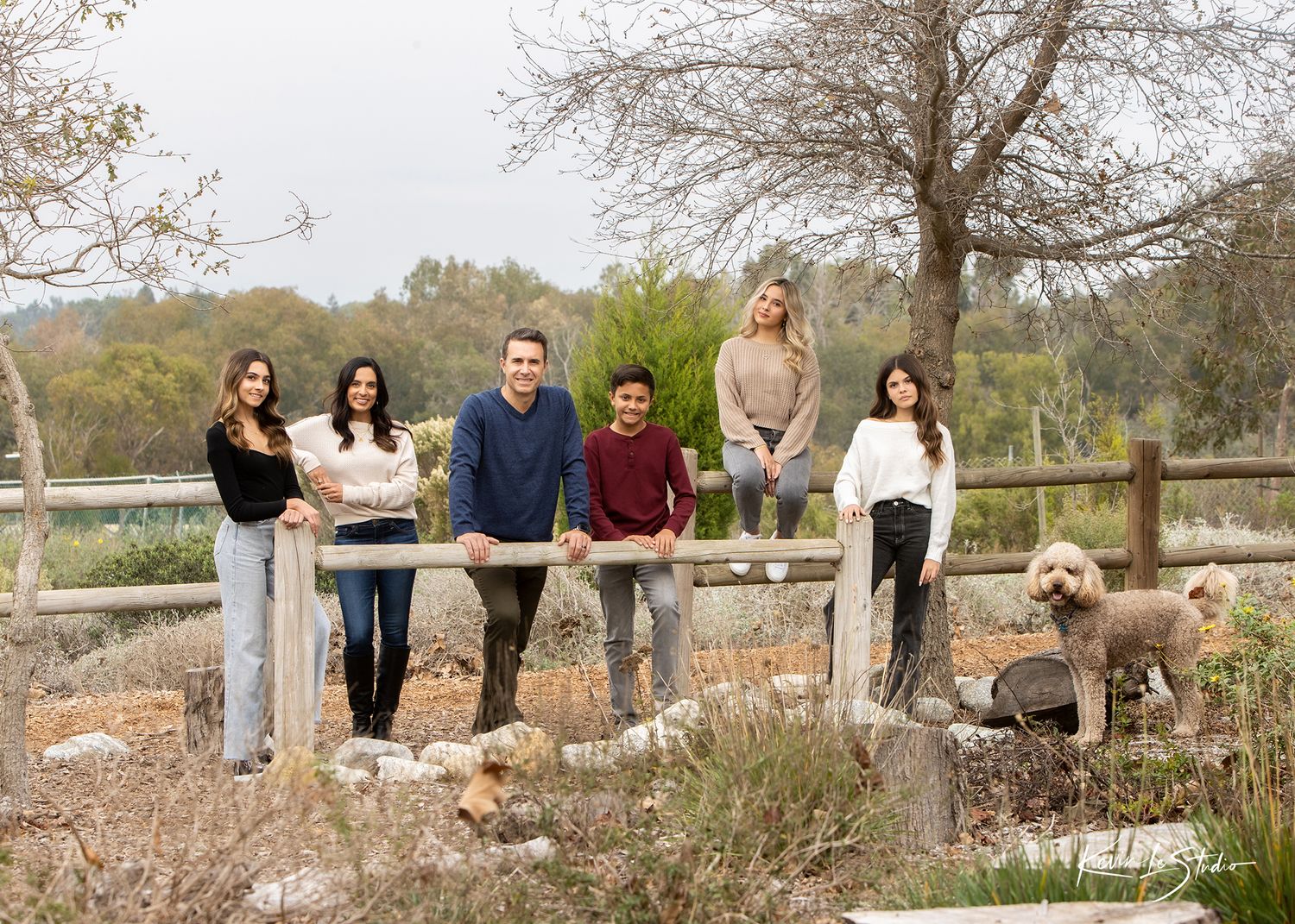 Family of six poses with a golden doodle dog by a rustic wooden fence in an outdoor nature setting.