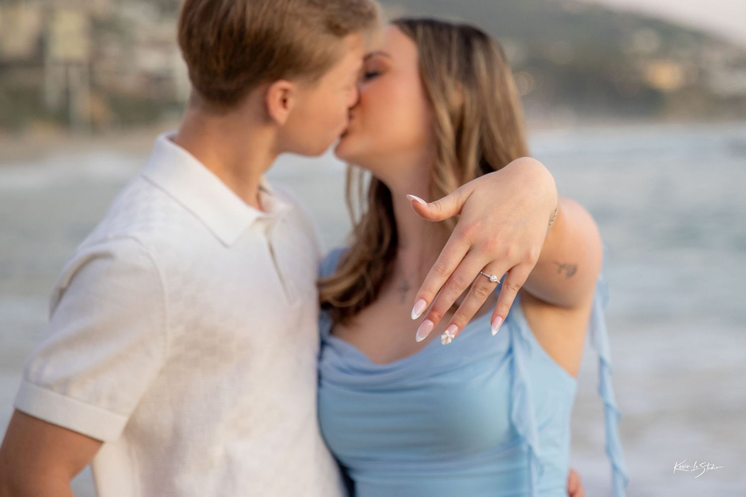 Newly engaged couple kissing on beach, woman displaying diamond engagement ring.