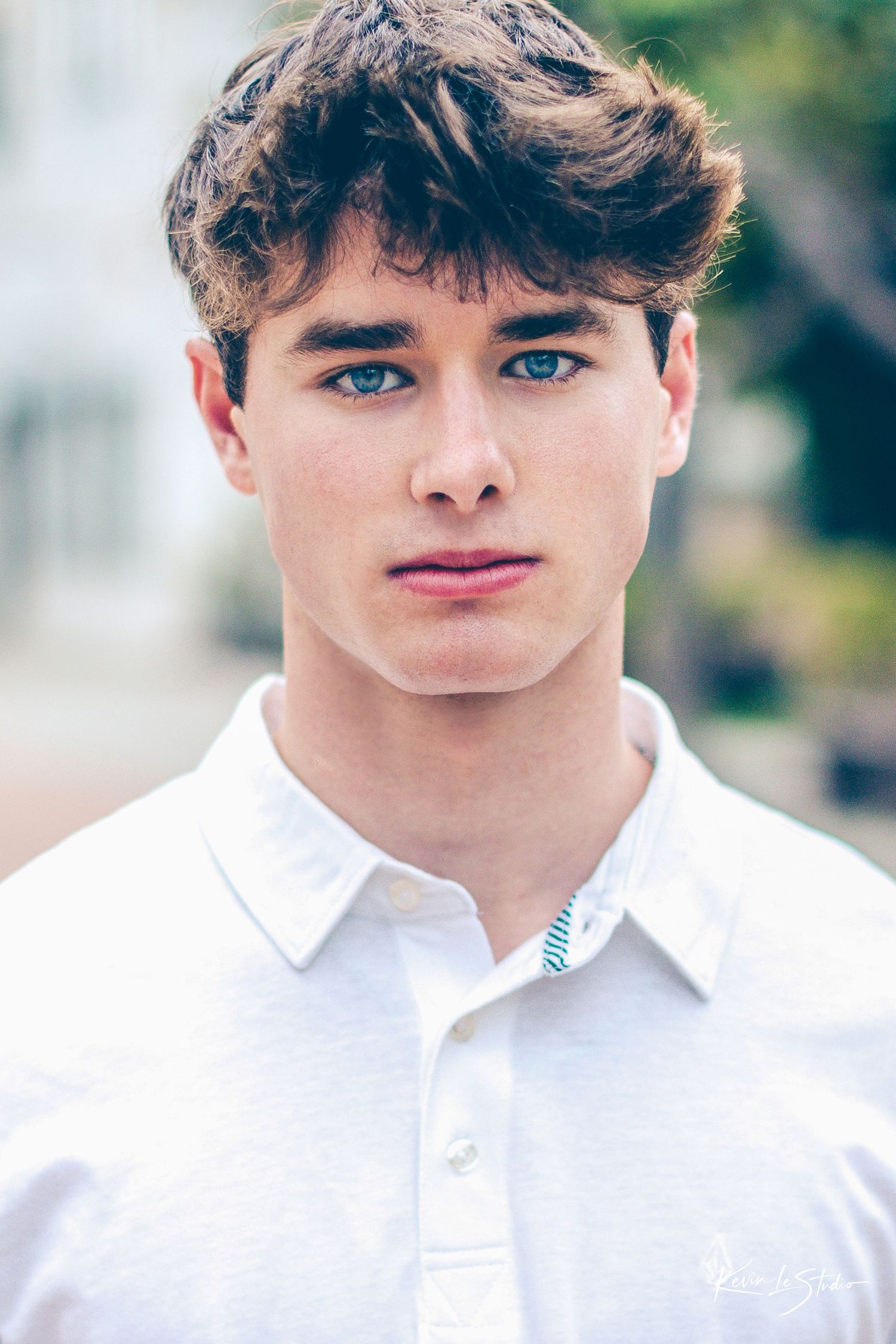 Close-up portrait of a young man with blue eyes and dark curly hair wearing a white polo shirt.