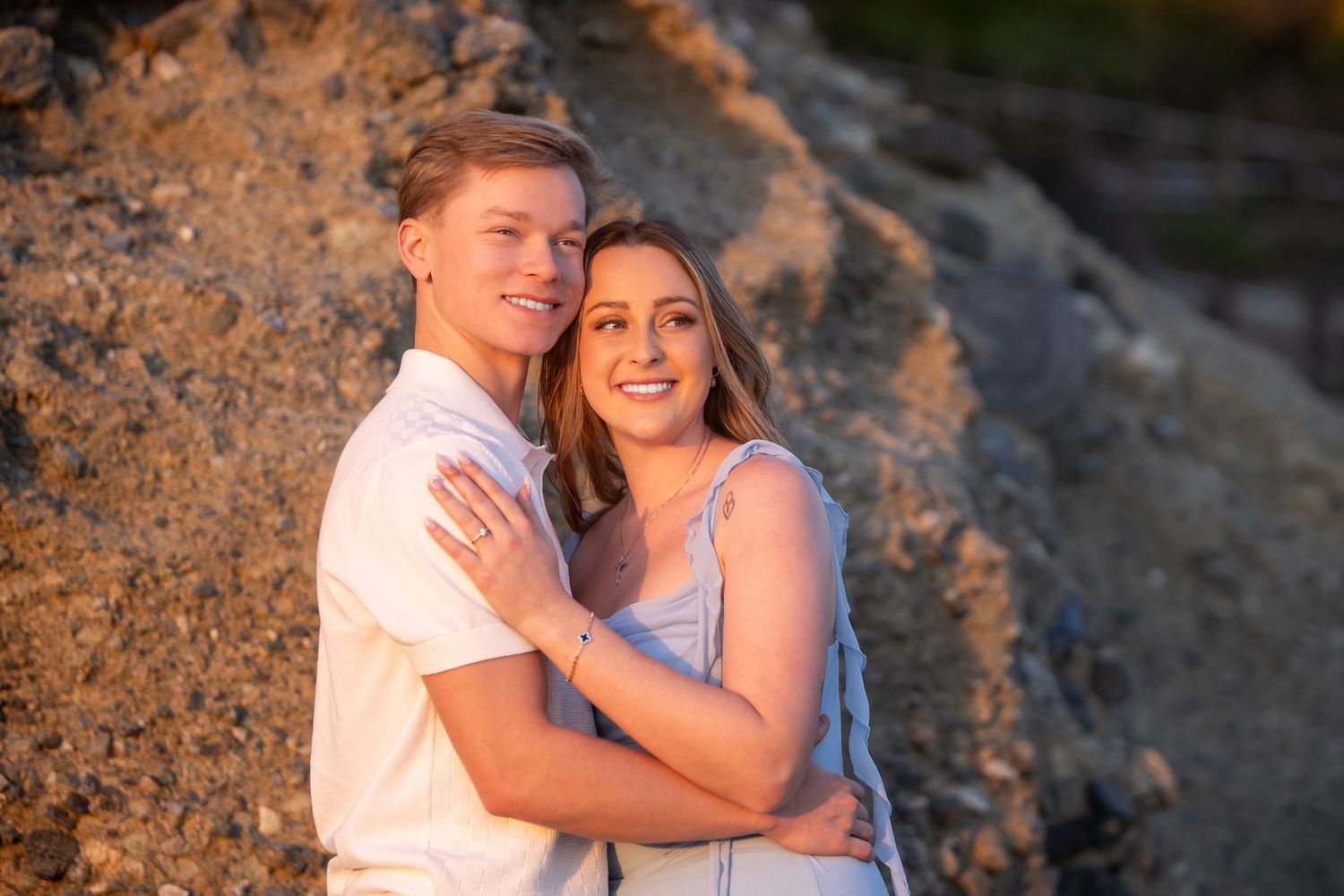 Smiling couple embracing in front of golden sunlit rocks during beach engagement session.