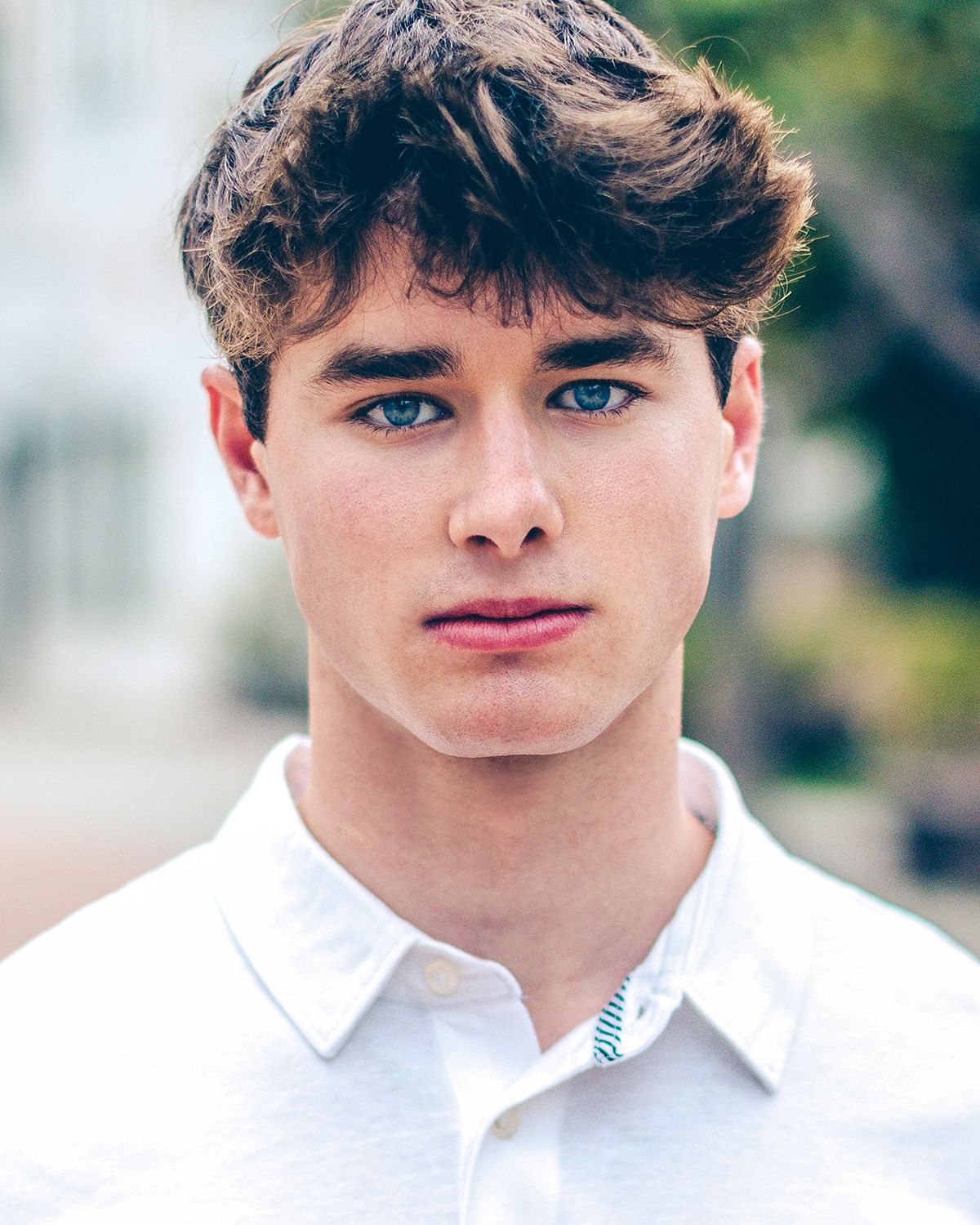 Young man with blue eyes and brown hair in white shirt posing outdoors.