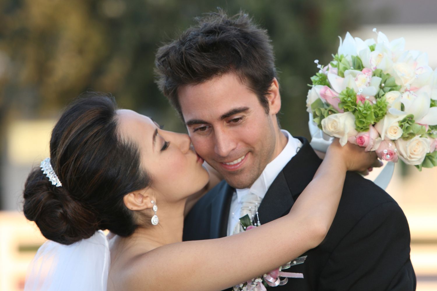 Joyful bride kissing groom on the cheek while holding a pink and white wedding bouquet outdoors.