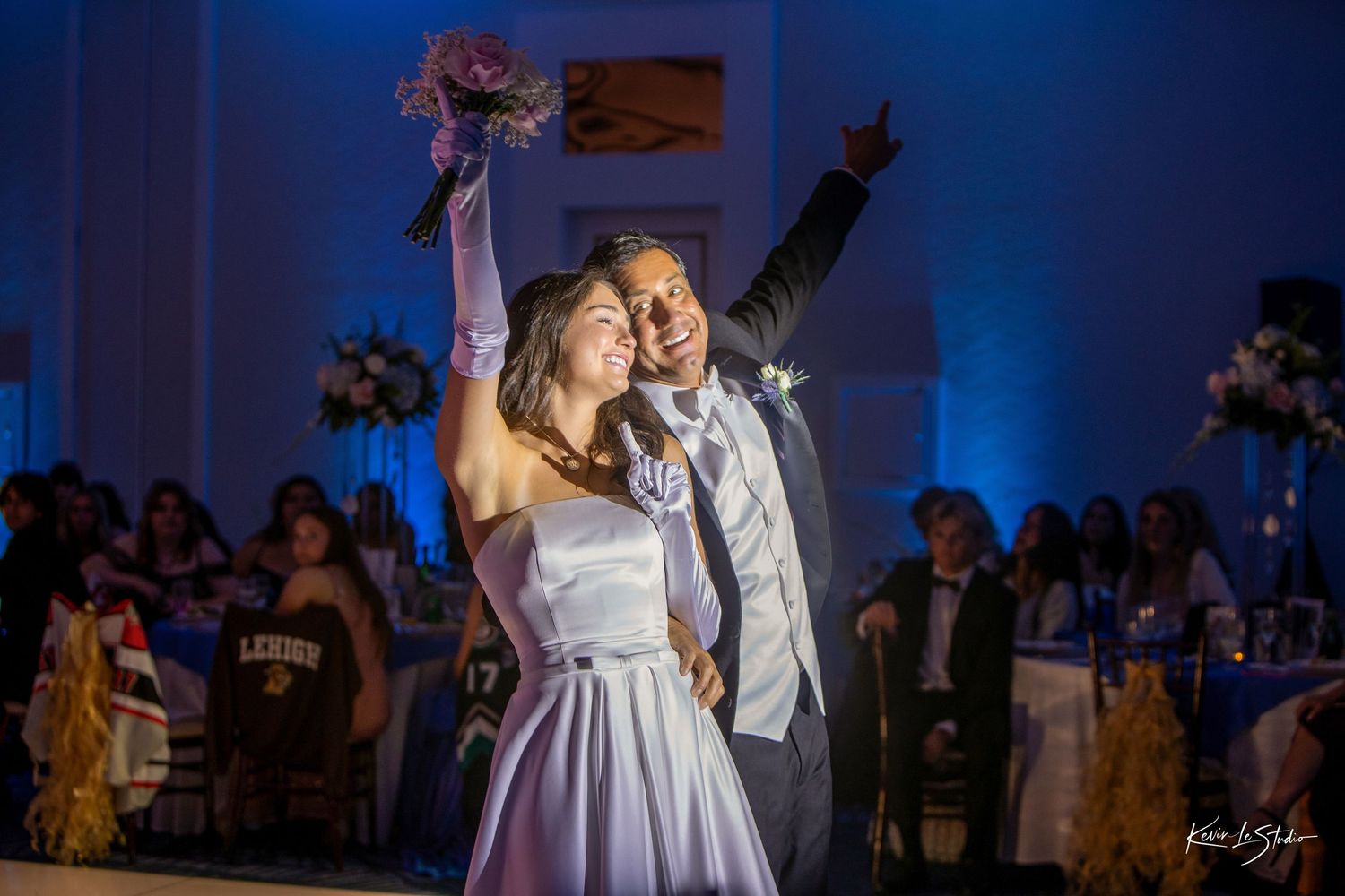 Joyful newlyweds celebrate at wedding reception with blue lighting, bride holding bouquet aloft, groom pointing upward.