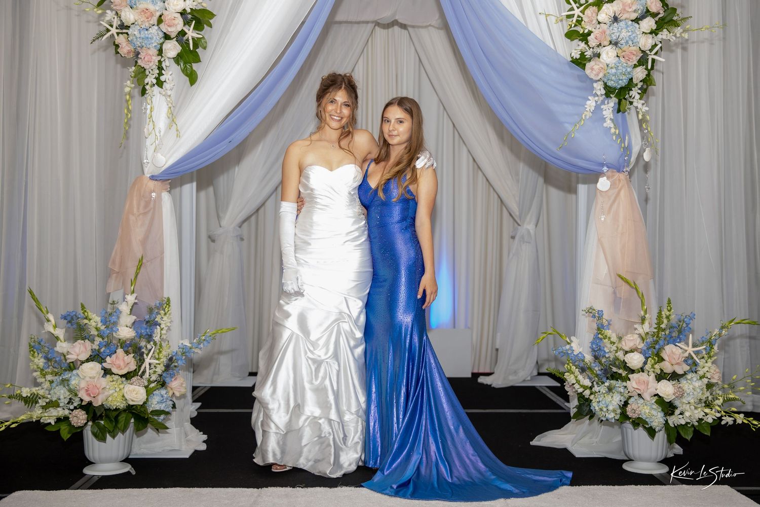Two women pose at a wedding, one in a white bridal gown and one in a royal blue satin gown, framed by floral decor.
