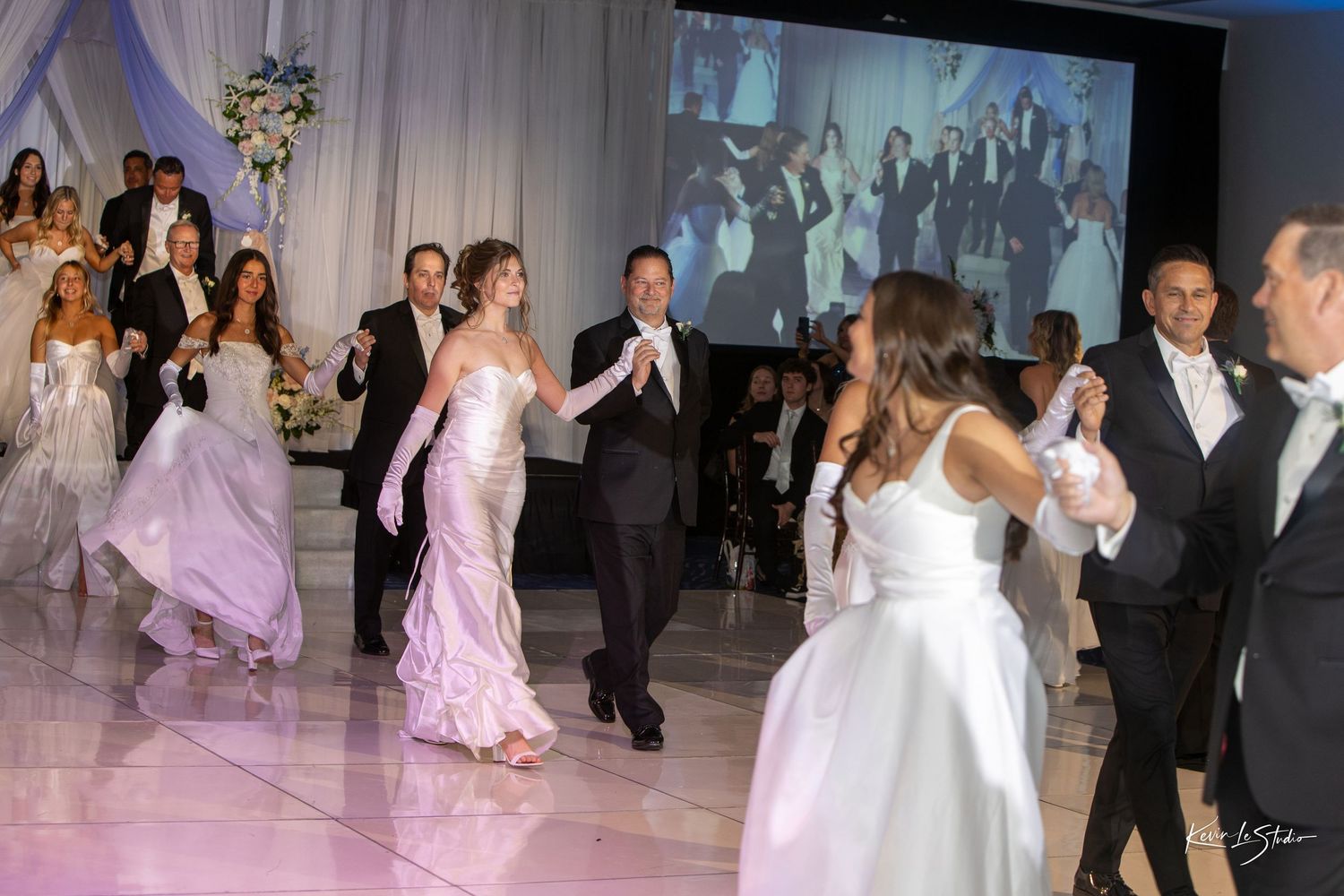 Elegantly dressed couples dance at a formal wedding reception with a projection screen displaying guests in the background.