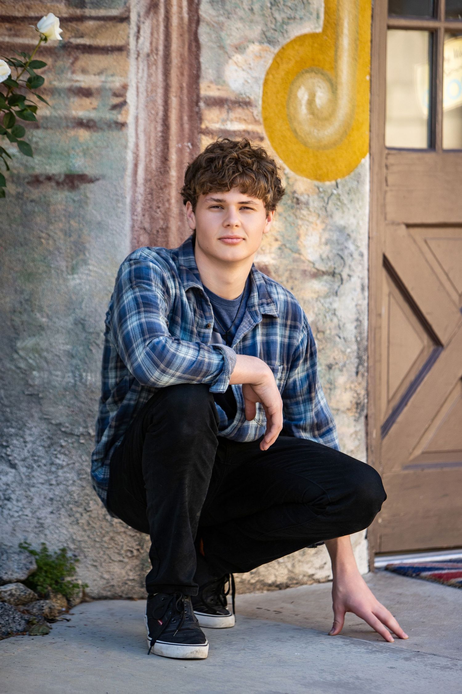 Teen boy in plaid shirt crouching outdoors near decorative wall with yellow spiral art.