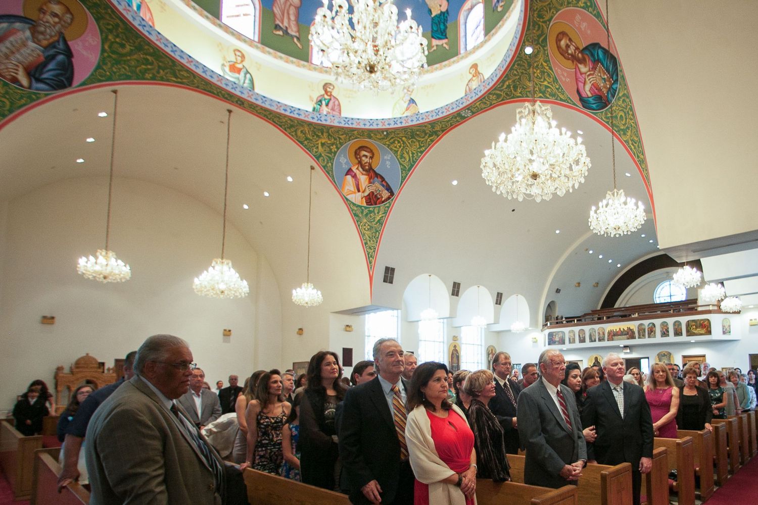 Congregation standing in ornate Orthodox church with domed ceiling, Byzantine icons, and elegant chandeliers during a service.