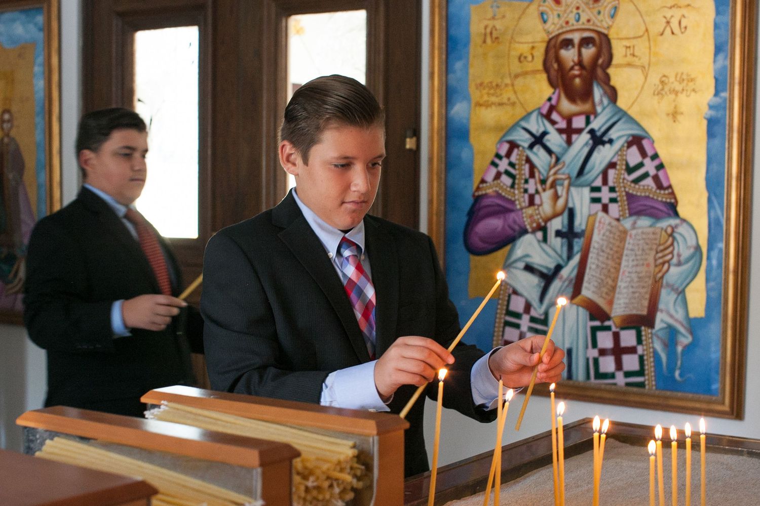 Two boys in suits lighting candles in an Orthodox Christian church with a Byzantine icon of Jesus Christ in the background.
