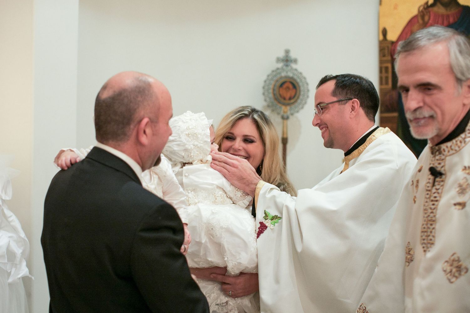 Parents and priest blessing infant in white christening gown during baptism ceremony.