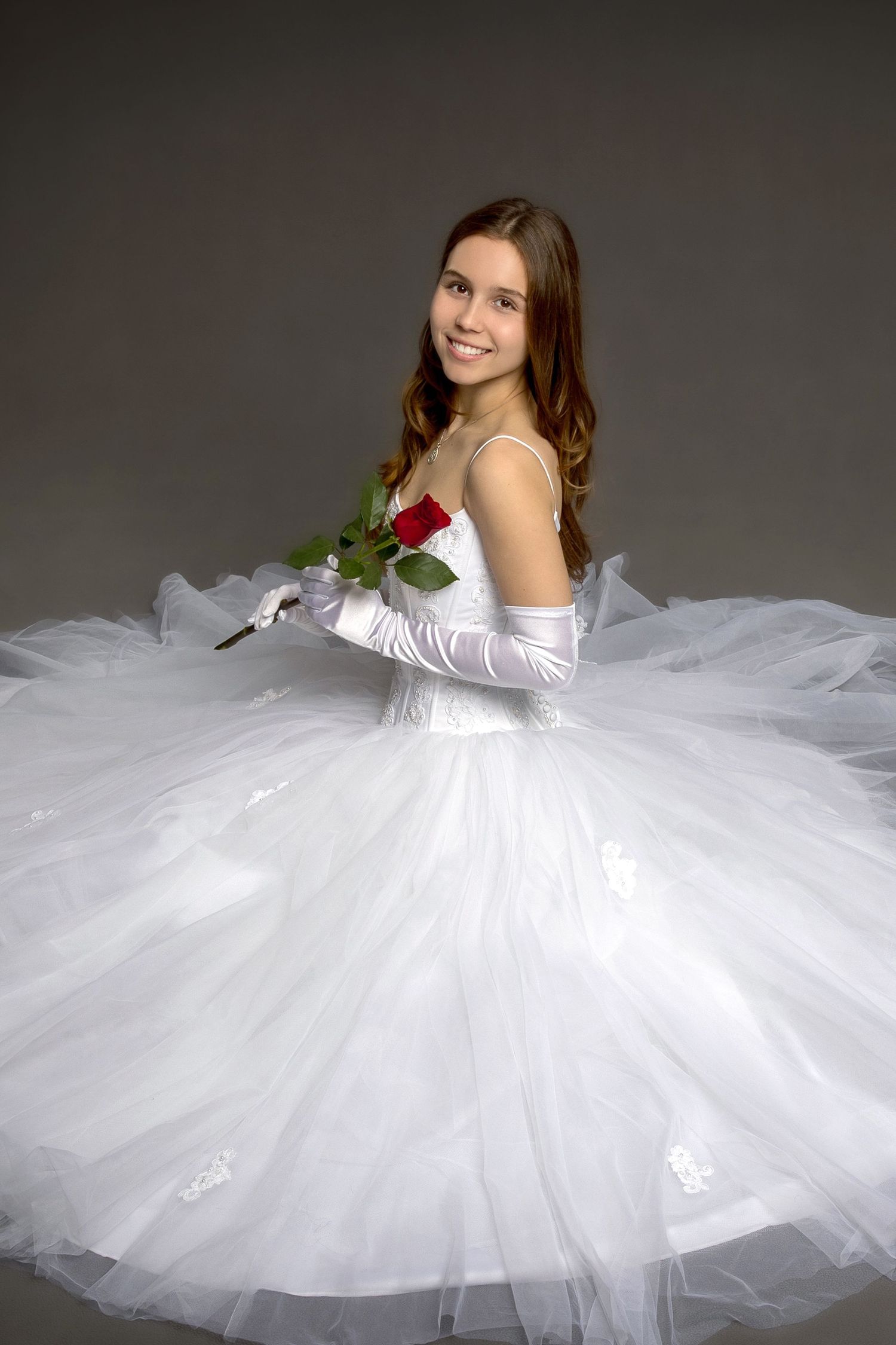 Young woman in white ball gown holding a red rose, smiling gracefully in a studio portrait.
