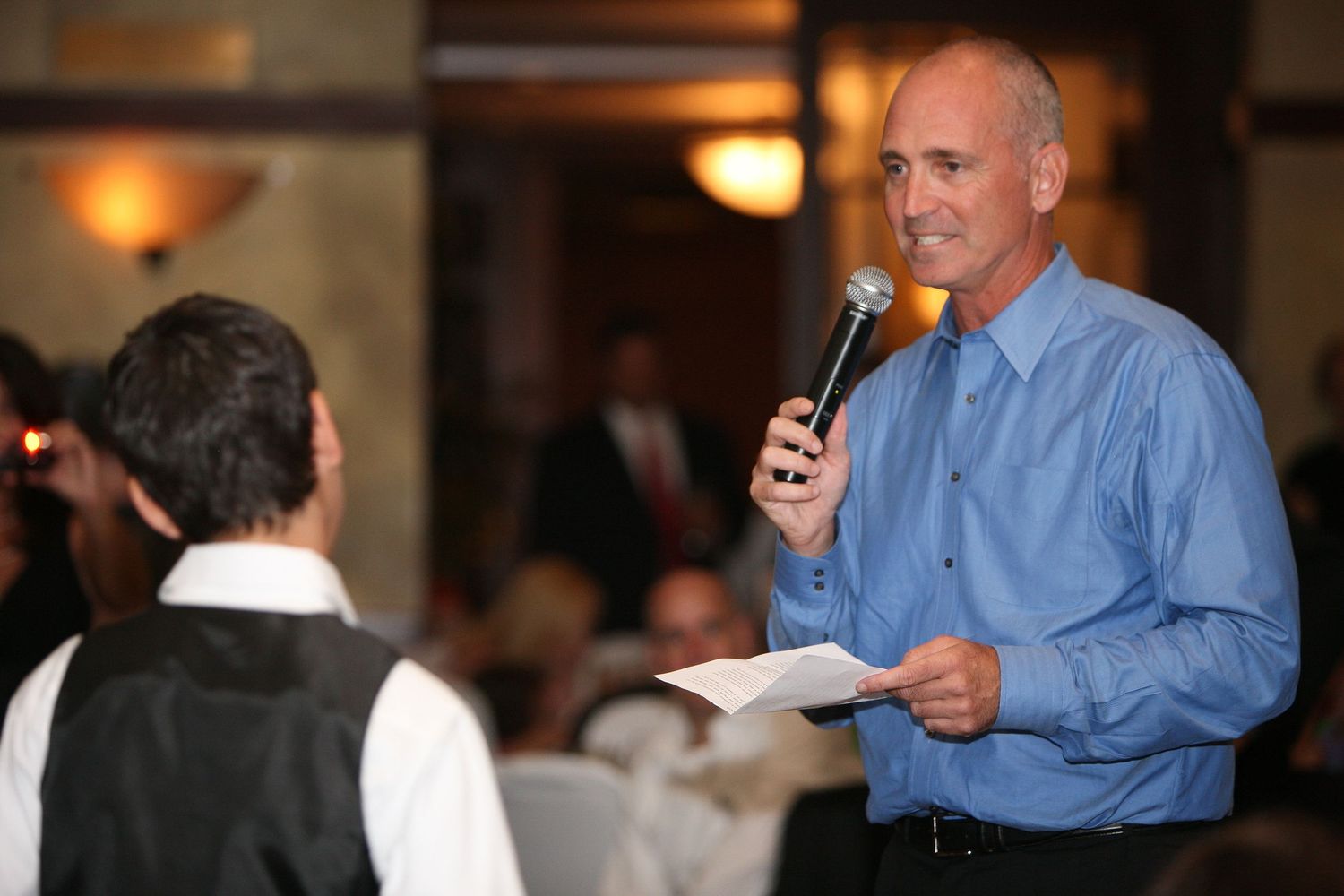 Man in blue shirt speaking into microphone at formal event, holding paper while addressing audience.