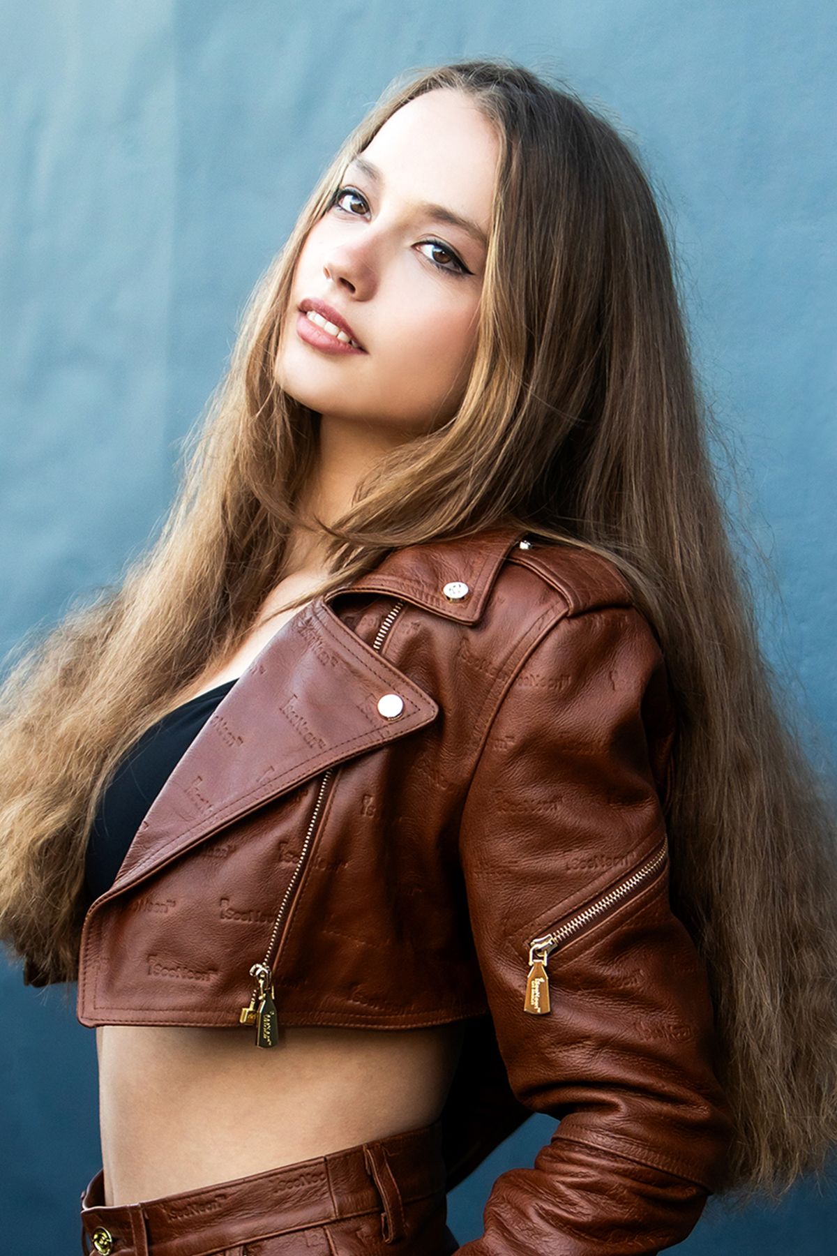 Young woman with long brown hair posing in a brown leather biker jacket against a blue wall.