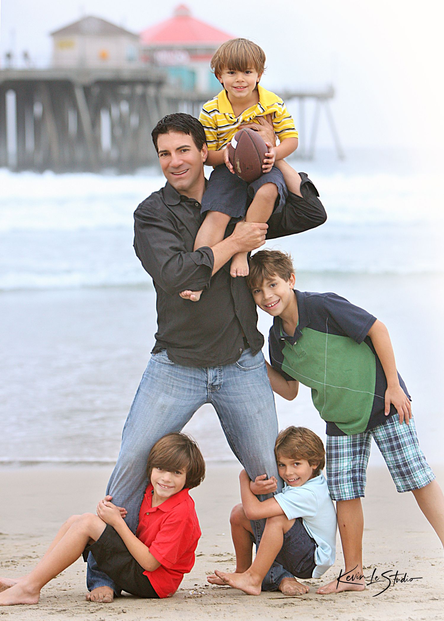 Father poses with four young boys on a sandy beach near a pier, holding a football in a fun family portrait.