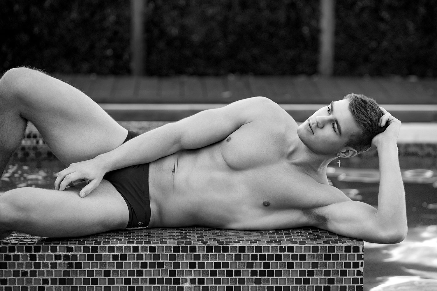 Black and white photo of a fit young man in swim briefs relaxing by a pool edge in bright sunlight.