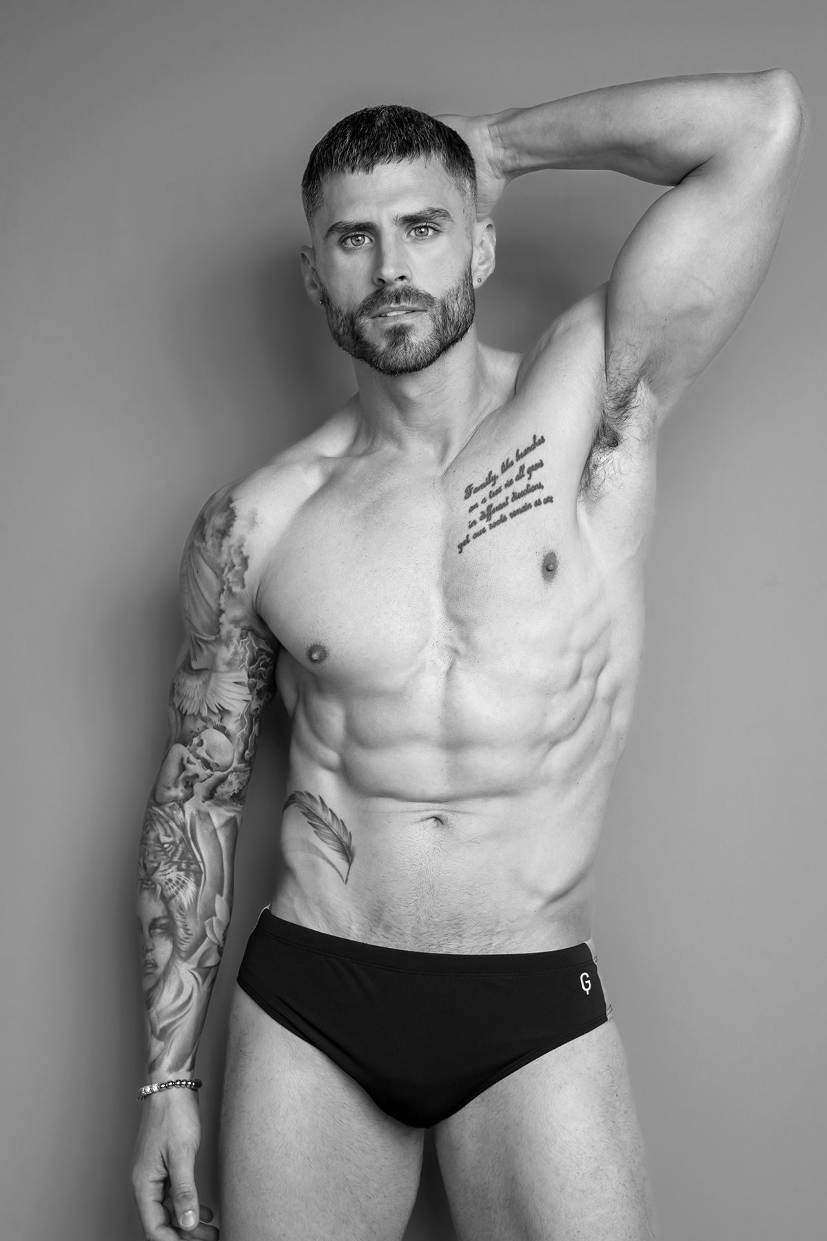 Black and white photo of a muscular tattooed man in swim briefs posing shirtless against a neutral background.