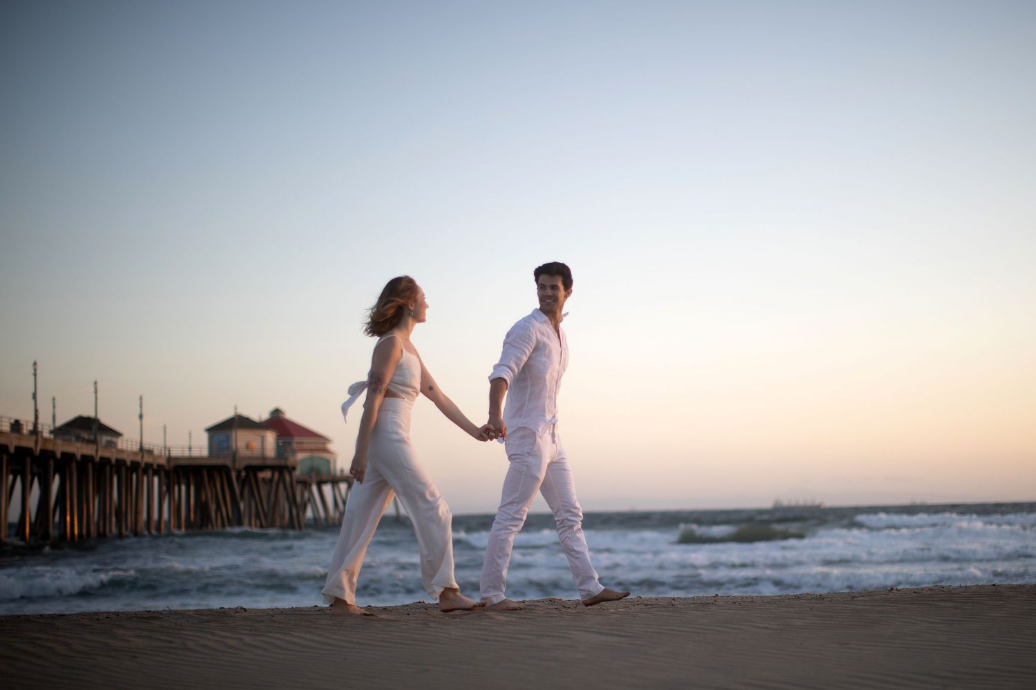 Couple in white outfits holding hands walking on beach at sunset near pier.
