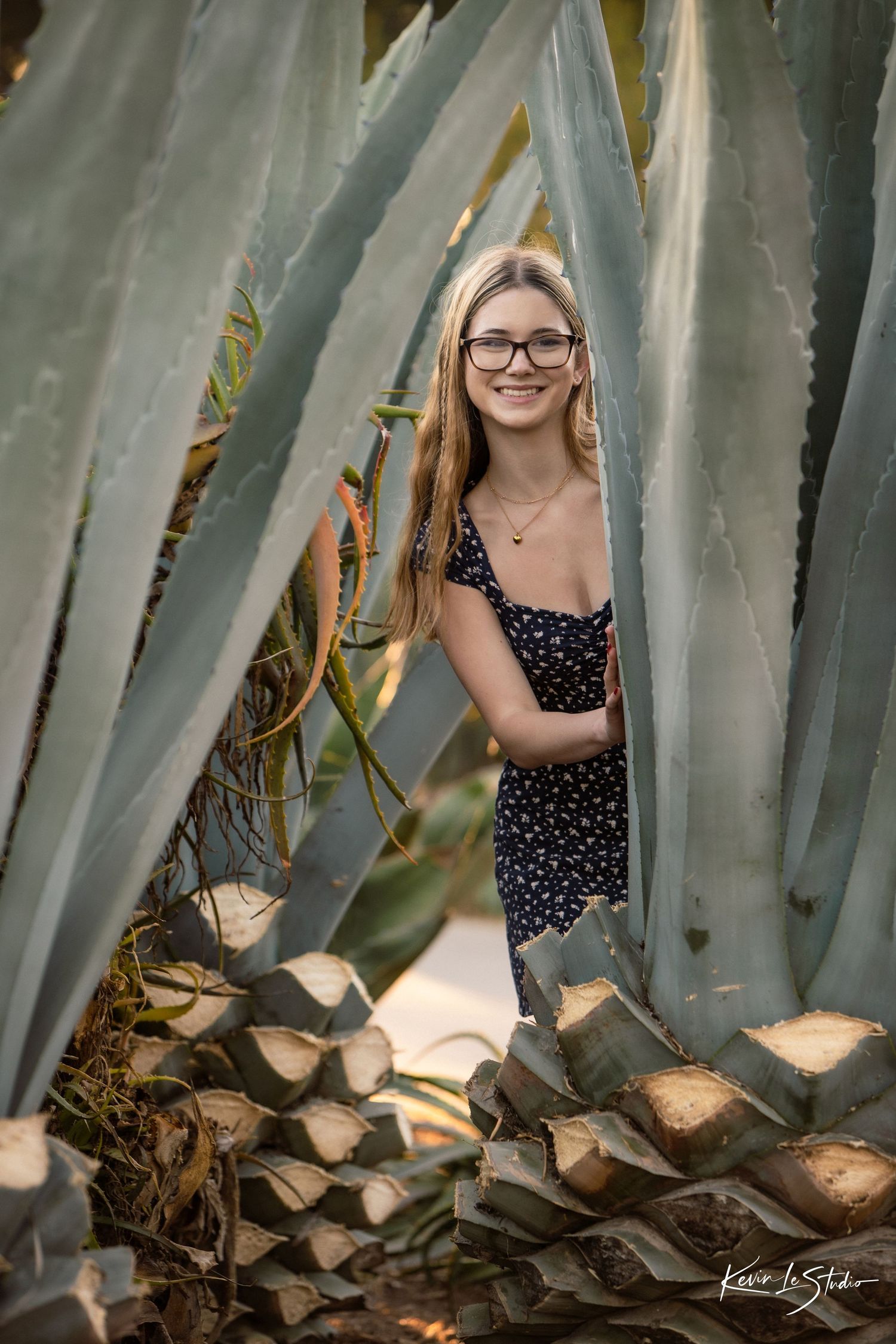Smiling teenage girl with glasses posing among large agave plants in a navy floral dress.