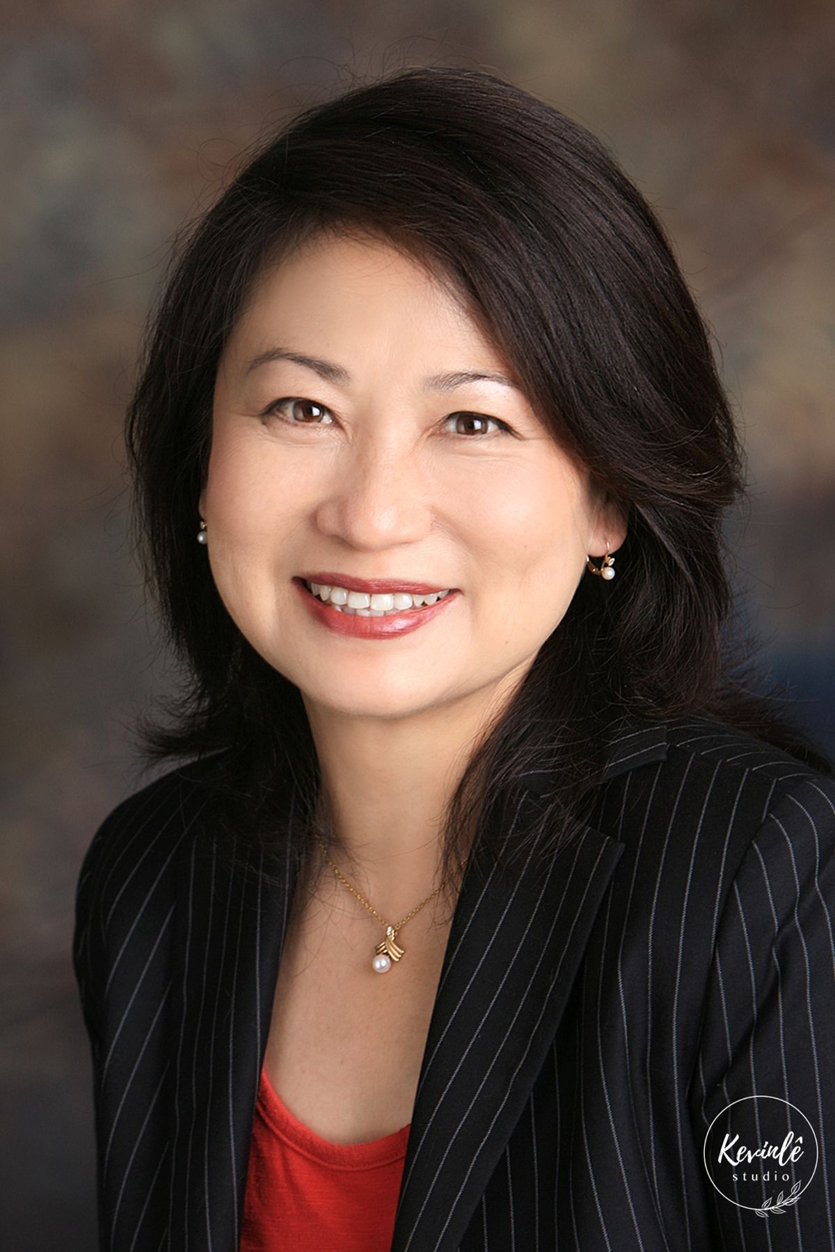 Professional headshot of a smiling Asian woman in a black pinstripe blazer with a pearl necklace.