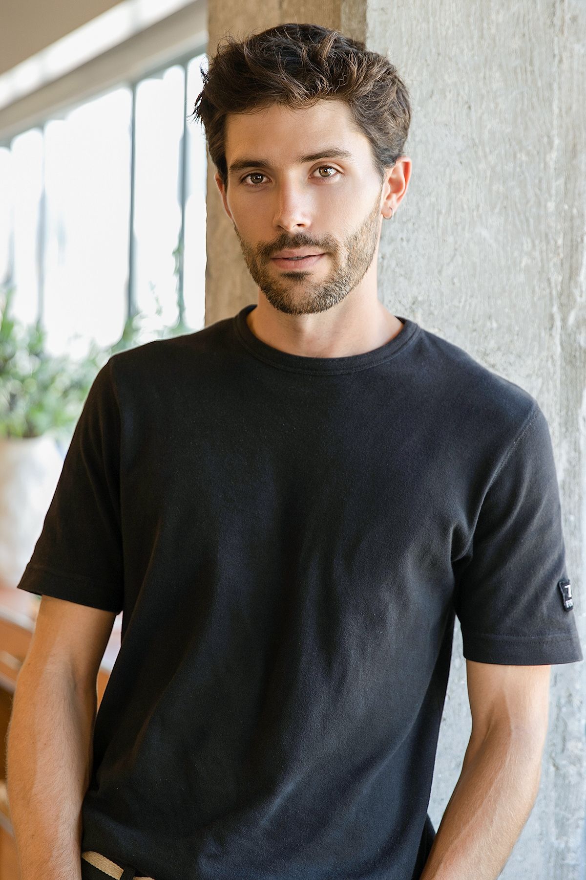 Bearded man with dark hair wearing a black crew-neck t-shirt, standing near a concrete pillar indoors.