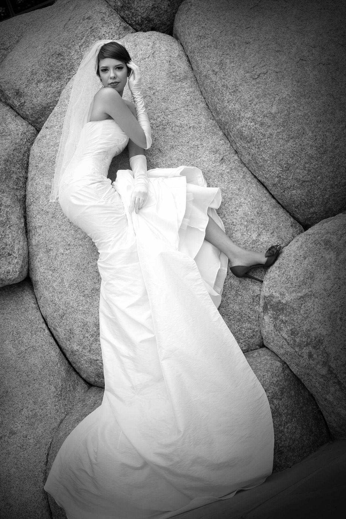 Bride in white wedding gown and veil poses gracefully on large rocks in a black and white portrait.