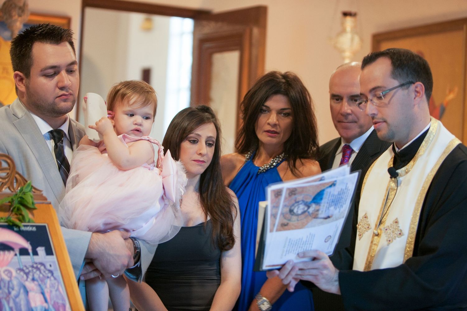 Family gathered for a baby baptism ceremony with a priest reading from a religious book in an elegant indoor setting.