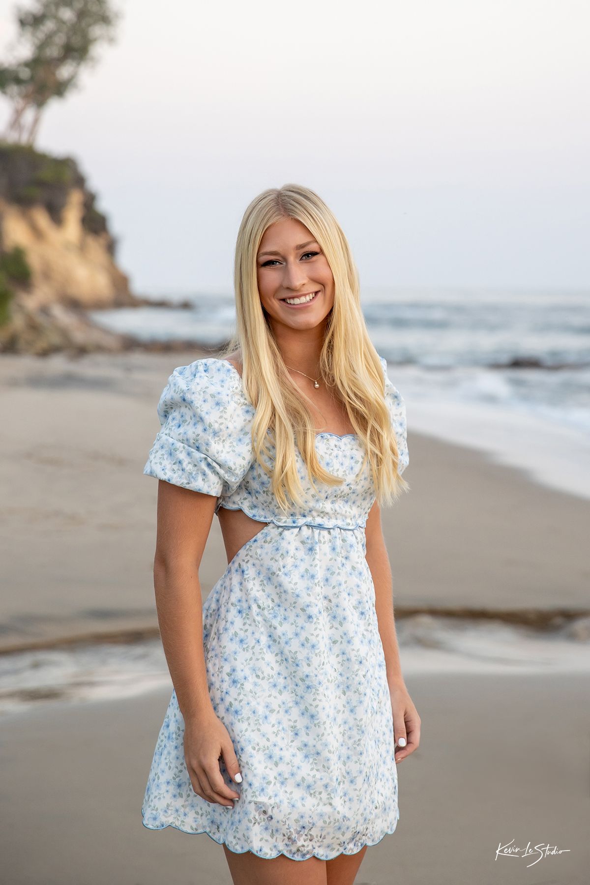 Blonde teen in white floral dress smiling on sandy beach at sunset.