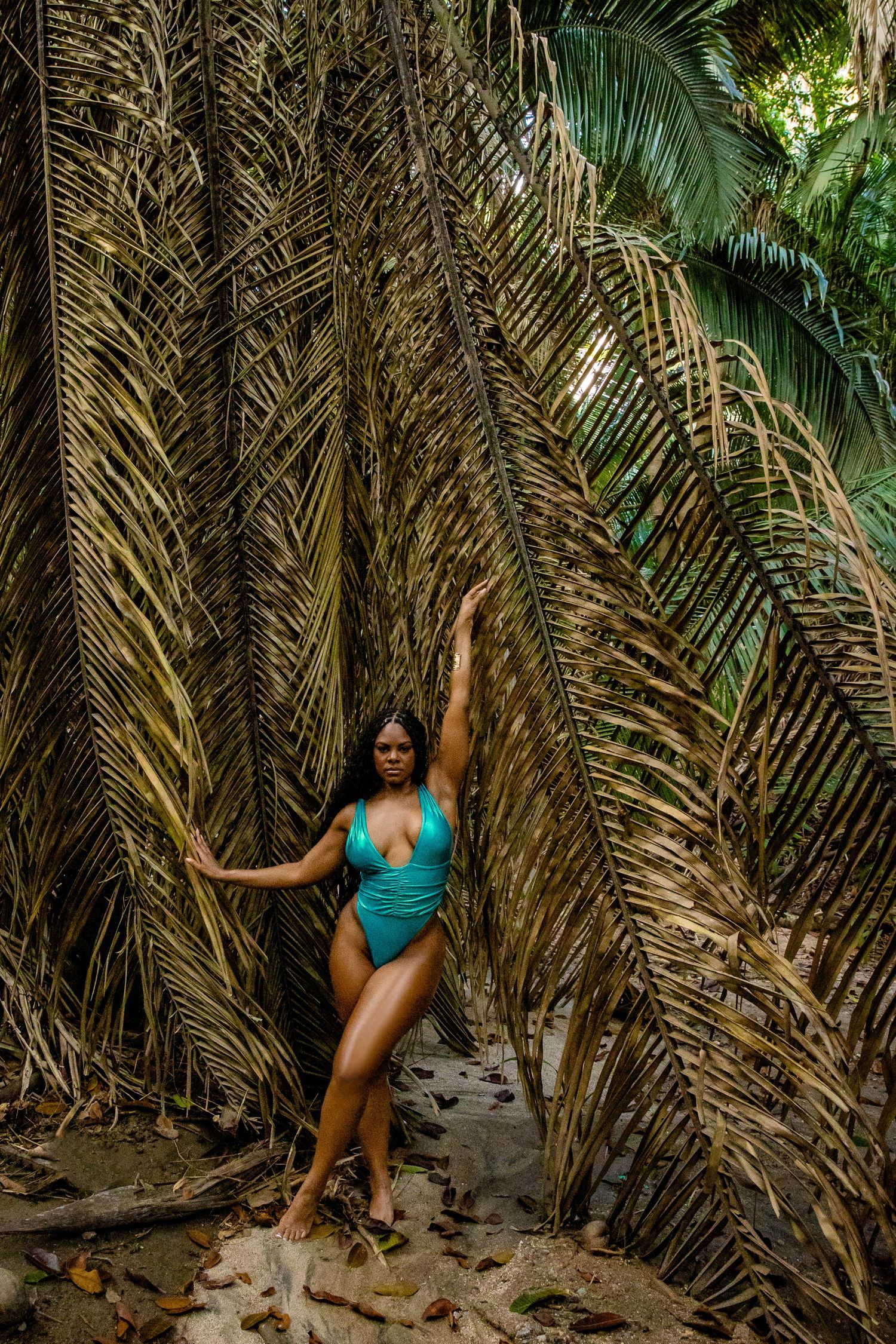 Woman in teal one-piece swimsuit posing among tropical palm fronds in a lush jungle.