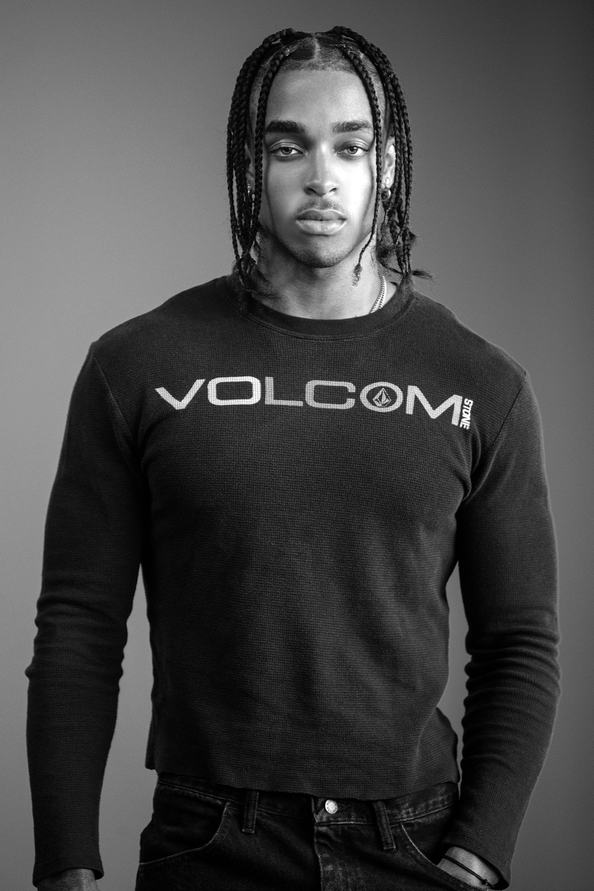 Young Black man with braids wearing Volcom sweatshirt poses in black and white studio portrait.