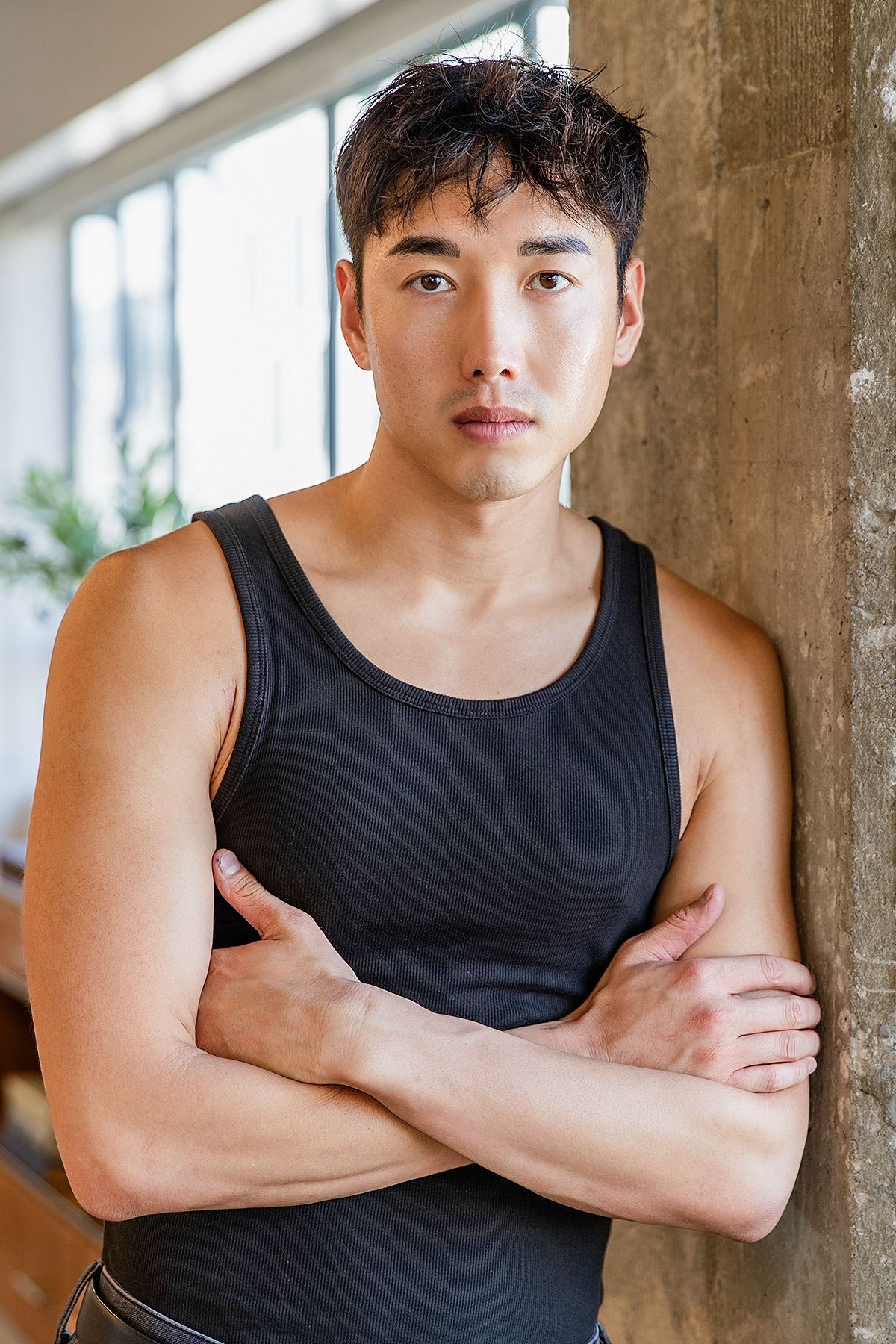 Muscular Asian man in black tank top with arms crossed leaning against concrete pillar indoors.