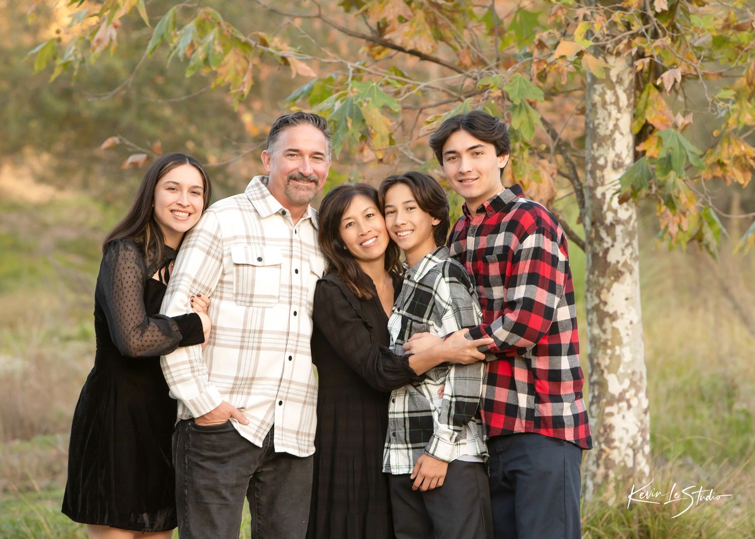 Family of five smiling together outdoors near a tree with autumn foliage in a professional portrait.