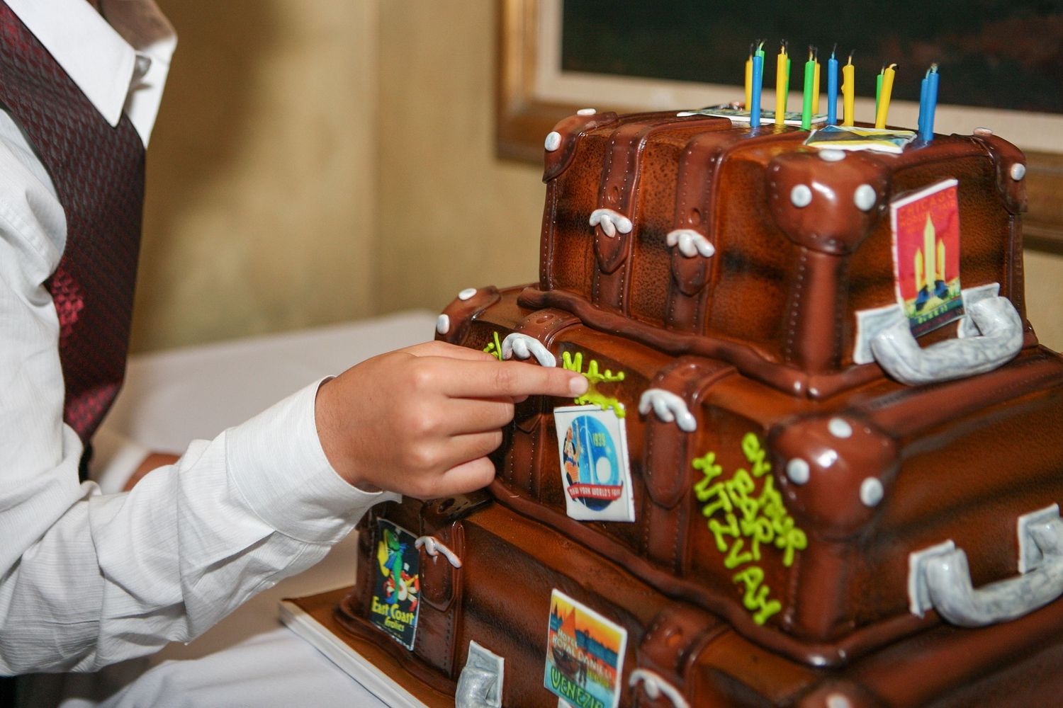 Tiered birthday cake decorated as vintage brown leather suitcases with travel stickers and colorful candles on top.