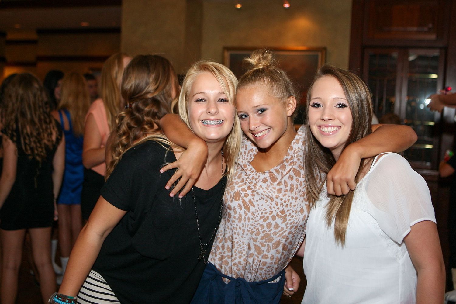 Three young women smiling and posing together at a social gathering, wearing casual outfits in a warmly lit venue.
