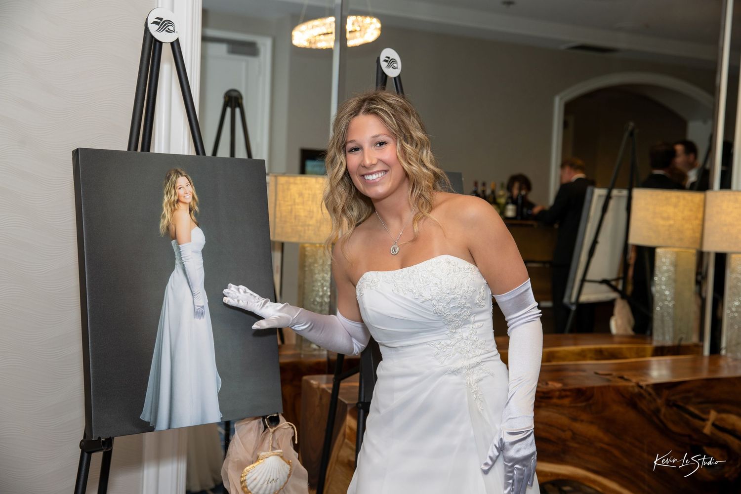 Bride in white strapless gown and gloves smiling beside a portrait painting of herself on an easel.